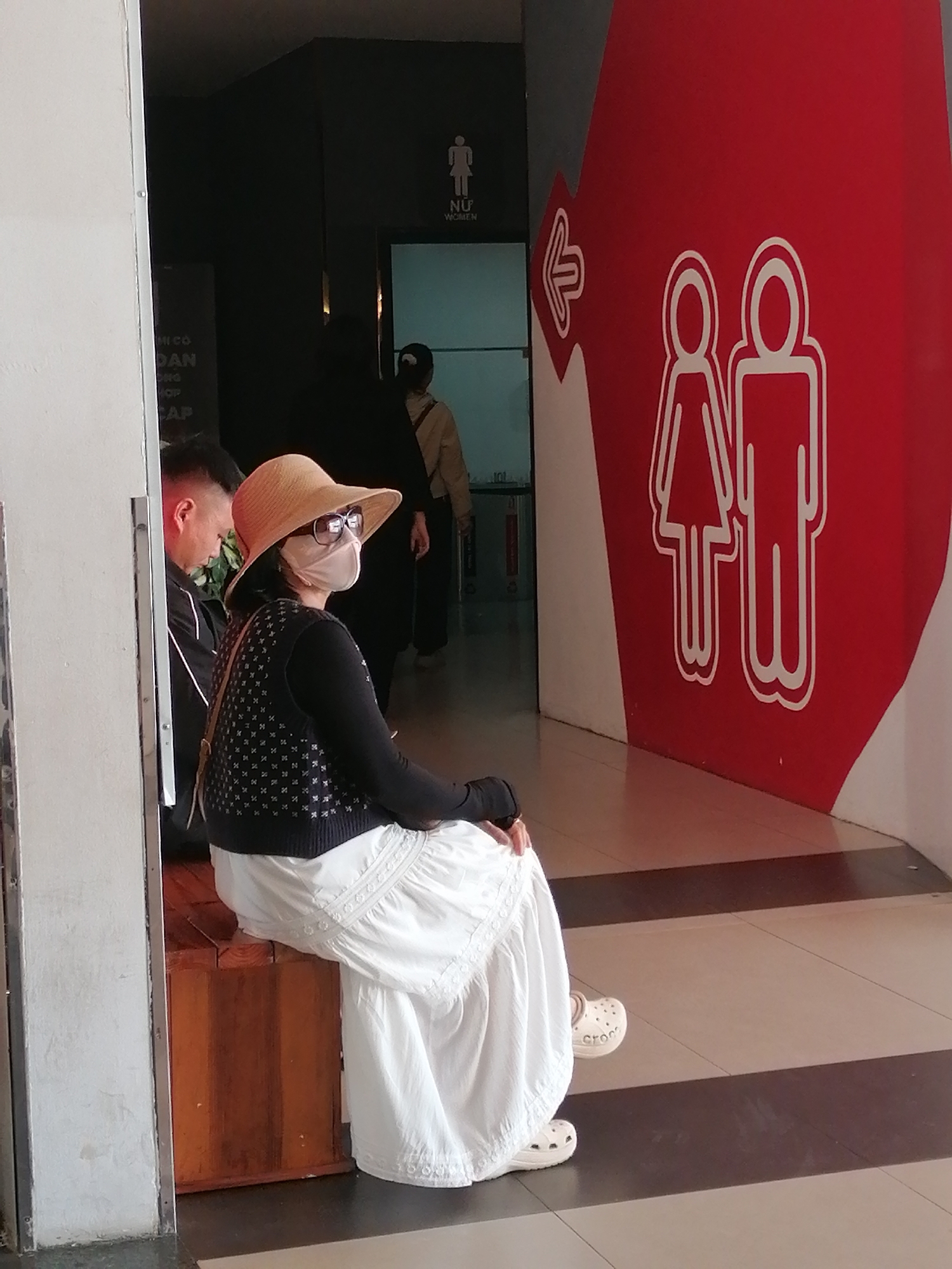 The image depicts an interior scene, possibly a public building, with a focus on a woman sitting on a wooden bench. She is wearing a light brown wide-brimmed hat, sunglasses, a pale pink face mask, a dark sleeveless vest with a white patterned design, and a long, flowing white skirt. She is also wearing white Crocs. To her left, a man is partially visible, wearing a black shirt. In the background, a sign with "NỮ WOMEN" and a gender symbol is visible above a doorway. To the right, a large red wall features stylized white icons of a man and a woman, indicating a restroom. An arrow pointing left suggests directions. The flooring is tiled with beige and dark brown stripes. The scene appears to be located in Da Lat, Vietnam, potentially near a tourist attraction or public facility.