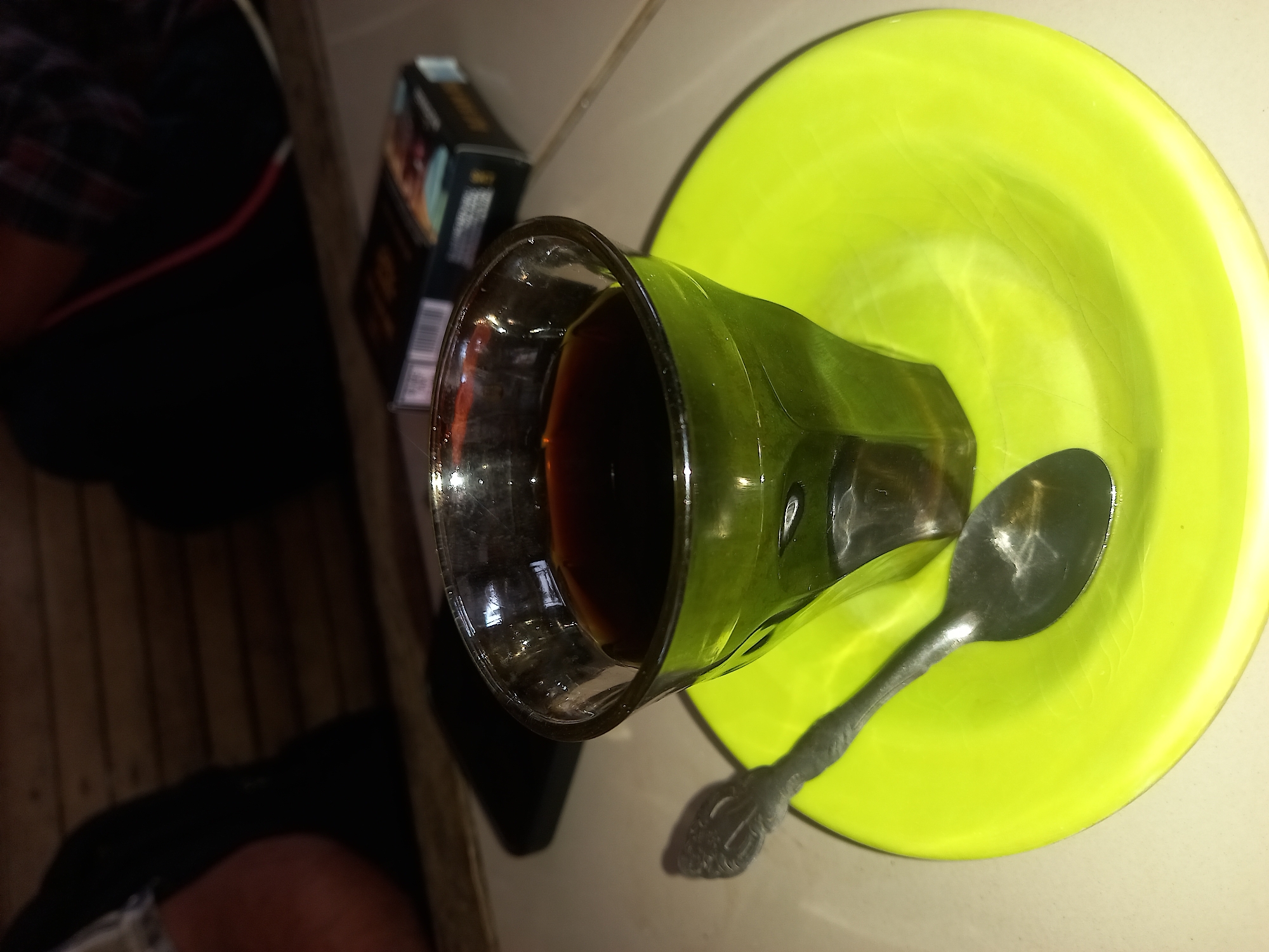 A close-up shot captures a glass of dark liquid, likely coffee, resting on a vibrant green saucer. A metal spoon lies on the saucer, angled to the left of the glass. In the background, a pack of cigarettes is visible, and hints of people and a wooden surface suggest an indoor setting. The image is from Keudejungkagajah, Indonesia.