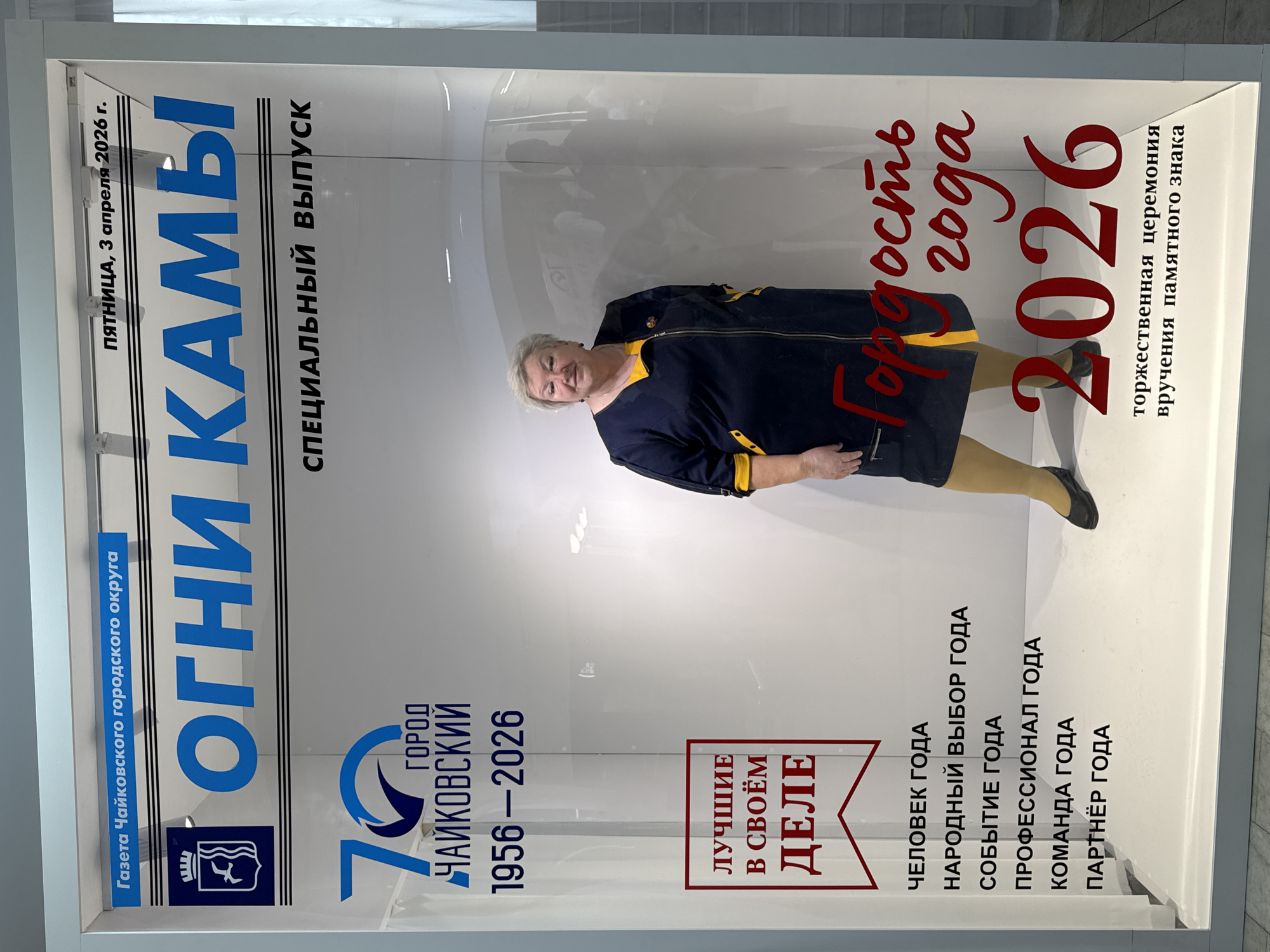 Here is a detailed description of the image:
**Scene:**
The image shows a woman standing in front of an advertisement panel behind glass. The panel appears to be promoting the city of Chaikovsky, Russia.
**People:**
The subject is a woman with short, light hair, wearing a navy blue jacket with yellow trim and leggings. She is standing and looking towards the camera. Her presence in front of the advertisement suggests a connection to it or possible promotion of it.
**Objects/Subjects:**
The main object is the advertisement panel, which is large and likely part of a display. Key elements of the advertisement include:
* Title: "ОГНИ КАМЫ" (Lights of Kama), likely the name of a newspaper or publication.
* Text: "Пятница, 3 апреля 2026 г." (Friday, April 3, 2026). "Специальный выпуск" (Special Edition). "70 город Чайковский 1956-2026" (70th city Chaikovsky 1956-2026)
* Slogan: "ЛУЧШИЕ В СВОЁМ ДЕЛЕ" (BEST IN THEIR PROFESSION).
* Categories: ЧЕЛОВЕК ГОДА (Person of the Year) НАРОДНЫЙ ВЫБОР ГОДА (People Choice of the Year), СОБЫТИЕ ГОДА (Event of the Year), ПРОФЕССИОНАЛ ГОДА (Professional of the Year), КОМАНДА ГОДА (Team of the Year) and ПАРТНЁР ГОДА (Partner of the Year)
* Slogan: "Гордость года 2026" (Pride of the Year 2026)
* Event: "торжественная церемония вручения памятного знака" (solemn ceremony of presenting a commemorative sign).
**Environment:**
The background is a lightly lit, modern interior, possibly in a public building or gallery where the advertisement is displayed. The surrounding area appears clean and well-maintained.
**Actions/Interactions:**
The woman is posing or standing still for the photograph, interacting with the advertisement panel by her presence.
**Additional details:**
The poster celebrates a year of pride in the city of Chaikovsky in 2026 and appears to be promoting a specific commemorative event. The inclusion of local newspaper and official symbols suggests this ad has official endorsement.