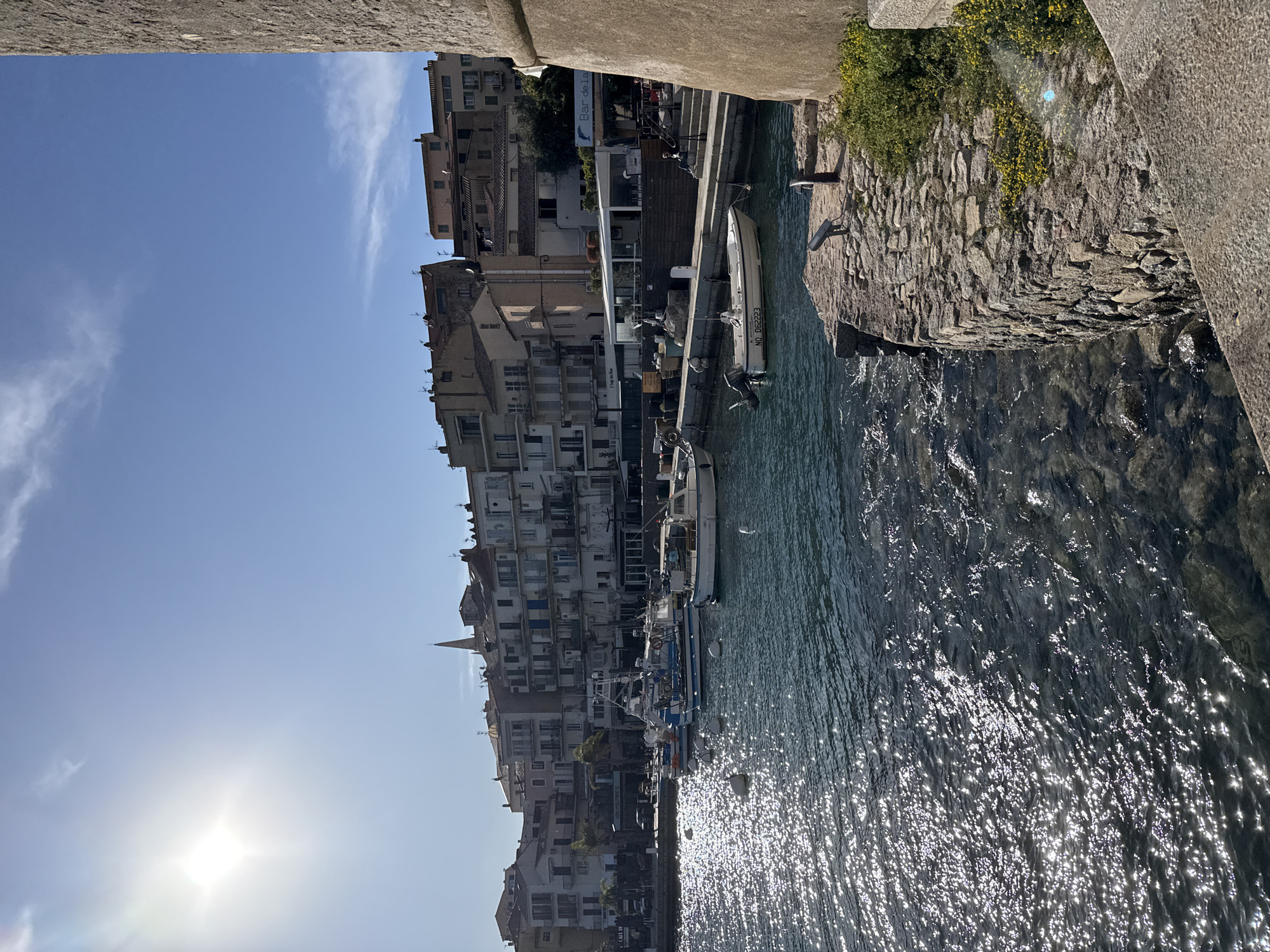 The image depicts a sunlit harbor scene in Calvi, France, viewed from a stone fortification. The upper left quadrant features a bright sun against a clear blue sky with minimal wispy clouds. The water in the foreground and midground is dark blue-green, intensely reflective with numerous sparkling glints from the sun, and exhibits ripples. Several white spherical buoys float on the water.
Along the far side of the harbor, a row of multi-story buildings with balconies, varying in color from white to light brown, lines the waterfront. Above these structures, a tall, pointed church spire is visible towards the left. At ground level along the promenade, several businesses are present, including a partially visible "Cafe Di" sign on the far left and a "Bar de La" sign further right. Palm trees are interspersed among the buildings.
Numerous boats are docked or moored in the harbor. On the left side, several blue and white fishing or leisure boats are visible, one featuring a prominent antenna and fishing equipment. In the mid-right, a small white motorboat with an outboard engine is moored, displaying the registration "NO D62223".
On the promenade, at least two individuals are present. One person is seated on stone steps near the "Bar de La" sign, facing away from the viewer. Another figure is discernible further left near the "Cafe Di" area.
The foreground and right side of the image are dominated by a massive, rough-hewn stone wall, part of a fortification, suggesting the viewpoint is from the historic Calvi Citadel. A lower stone retaining wall or dock extends along the water's edge, featuring some metal bollards and patches of green vegetation with small yellow flowers.