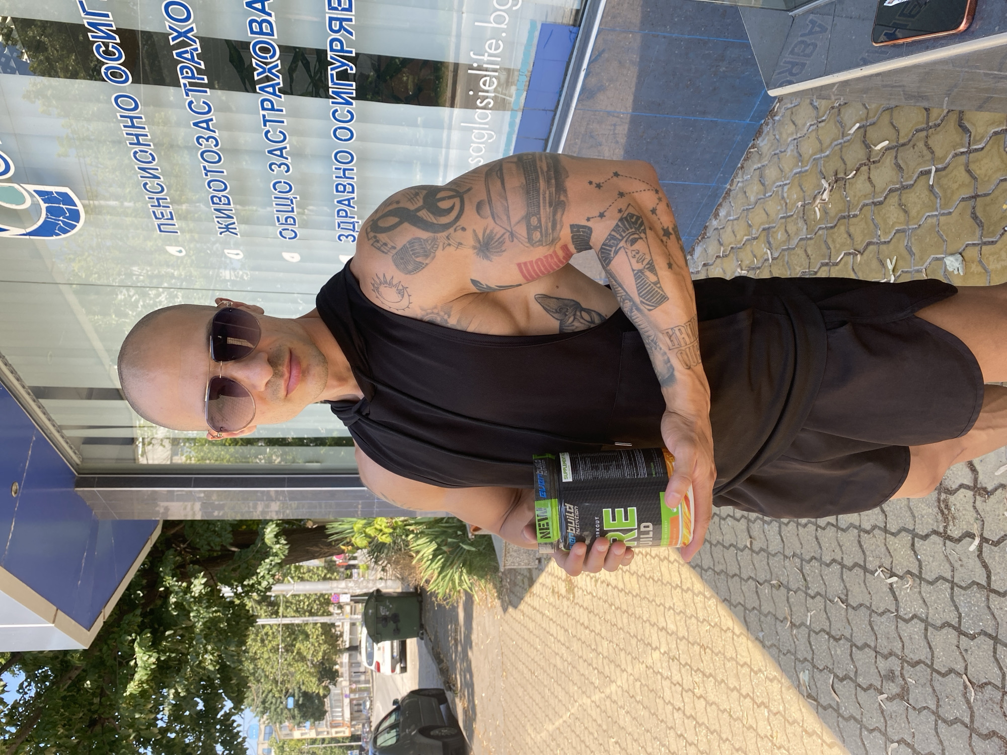 A bald man wearing sunglasses, a black sleeveless hoodie, and black shorts stands on a sidewalk in front of a building with a glass facade. He is holding a green and yellow container of dietary supplement. The building has blue lettering on its facade, which includes the text "PENSIONNO OSIGURYAVANE" and "ZDRAVNO OSIGURYAVANE". To the left of the building, a street with cars and trees is visible. The sidewalk is paved with yellow interlocking bricks. The background shows a street scene with trees, cars, and a green trash can. The man has tattoos on his arms. The overall scene appears to be outdoors in a city environment. Based on the Cyrillic text visible on the building's facade, the location is likely in Pleven, Bulgaria, as "osiguryavane" translates to "insurance" and these are common types of insurance offered in Bulgaria.