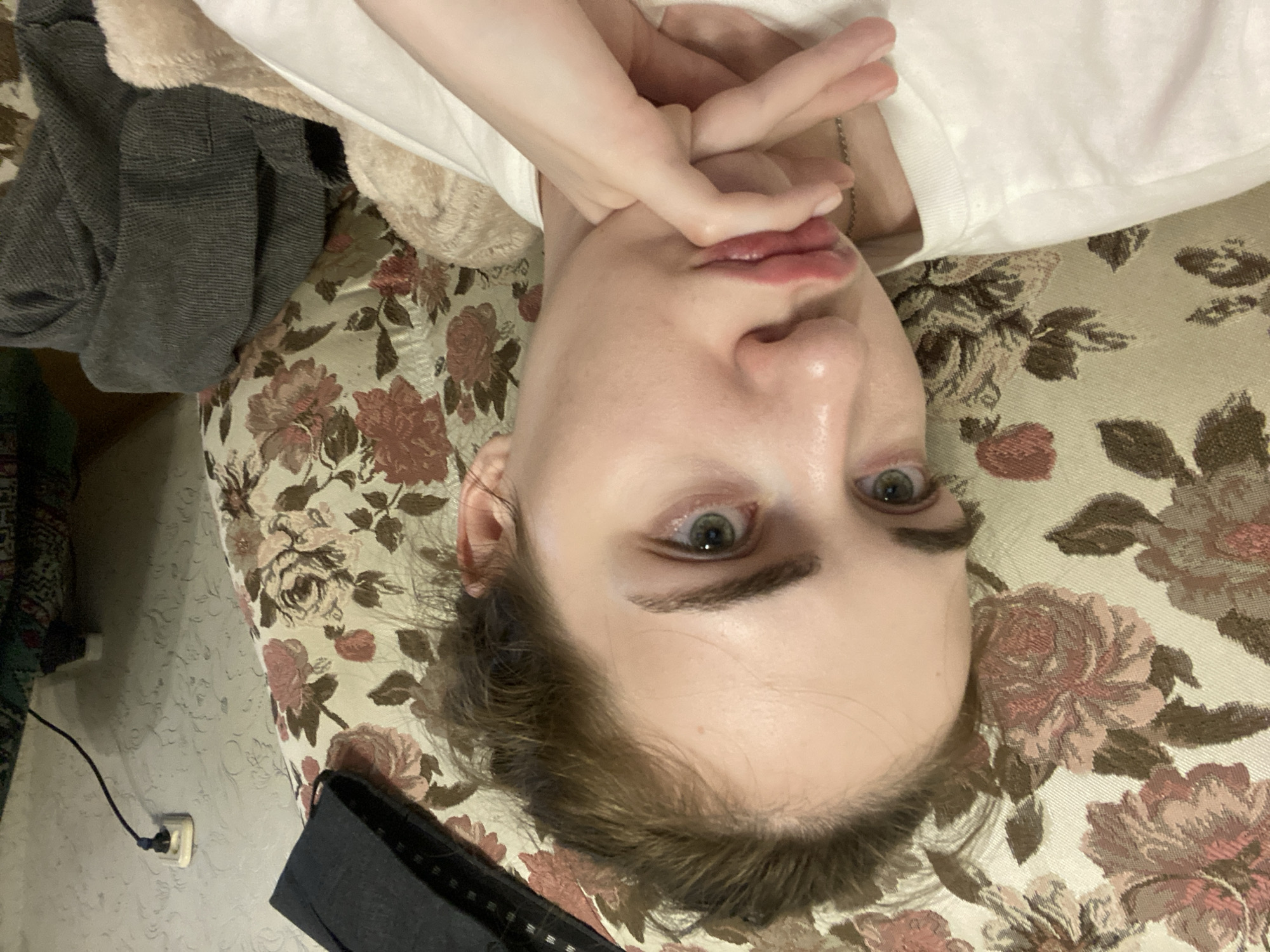 The image shows a person lying on a floral-patterned couch or bed. The person's head is tilted upwards and slightly to the left, with their hands crossed over their face, partially obscuring their features. The person is wearing a white t-shirt. A grey garment is visible on the left side of the frame, possibly clothing or a blanket. The background features a floral fabric with pink and brown roses.
On the left side of the image, a white electrical outlet with a black cord plugged into it is visible on a textured wall. The overall lighting suggests an indoor setting, possibly during the day. The image is oriented vertically, with the person's head at the top and their feet at the bottom if the image were rotated. The person's facial expression is not clearly discernible due to their hands covering their face, but their eyes are open and looking towards the camera. There is no visible text in the image. The location context of Zavodoukovsk, Russia, does not provide specific visual cues within this particular image.