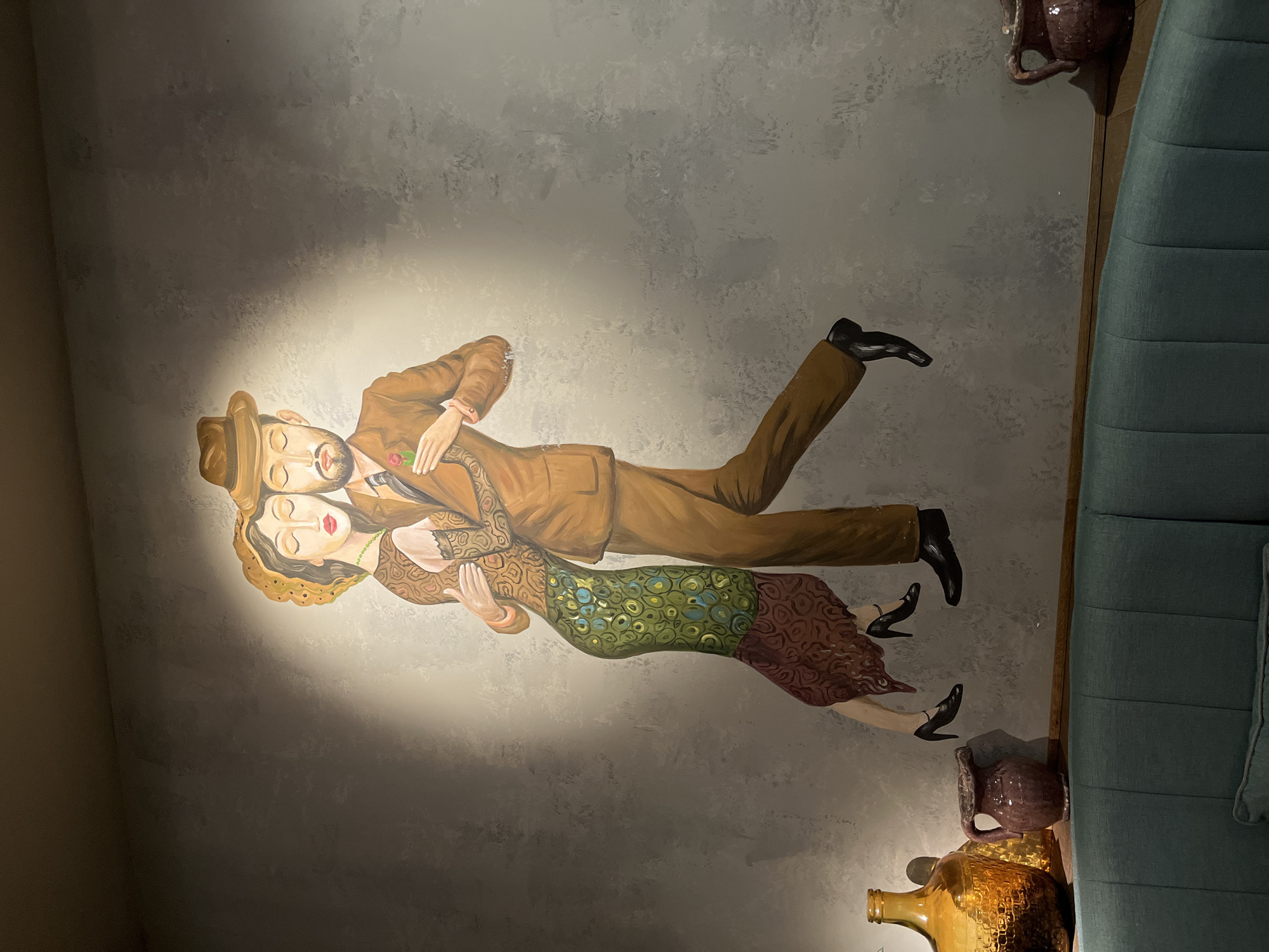 A mural depicting a man and a woman dancing is painted on a gray, textured wall in a dimly lit room. The man wears a brown suit and a fedora hat and holds a red rose. The woman wears a patterned dress and a hat. They are illuminated by a spotlight. In the foreground, a teal upholstered bench is visible, with a wooden shelf above it holding two decorative vases and a large amber glass bottle. The setting appears to be a restaurant or bar in Khimki, Russia.