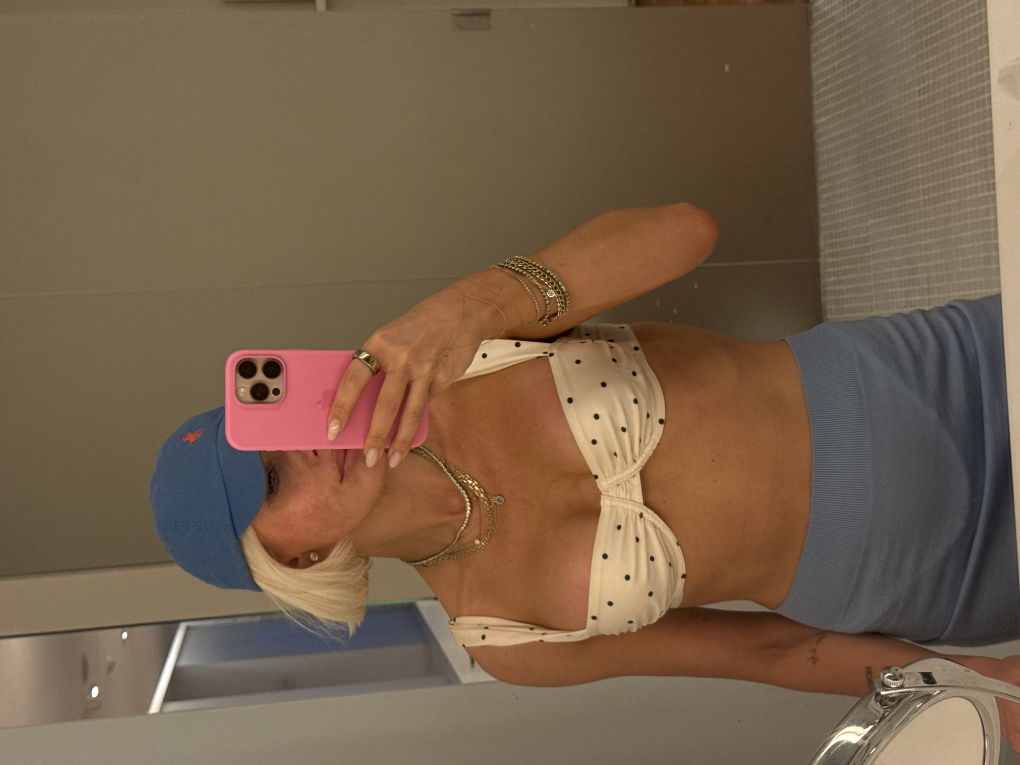 A blonde woman is taking a selfie in a bathroom. She is wearing a blue Ralph Lauren baseball cap, a white polka dot bikini top, and a blue skirt. She is holding a pink iPhone in her left hand. She is wearing several bracelets on her left wrist and a gold chain around her neck. The bathroom has light grey walls and a white tile wall. The photo was taken in Miami Beach, United States of America.
