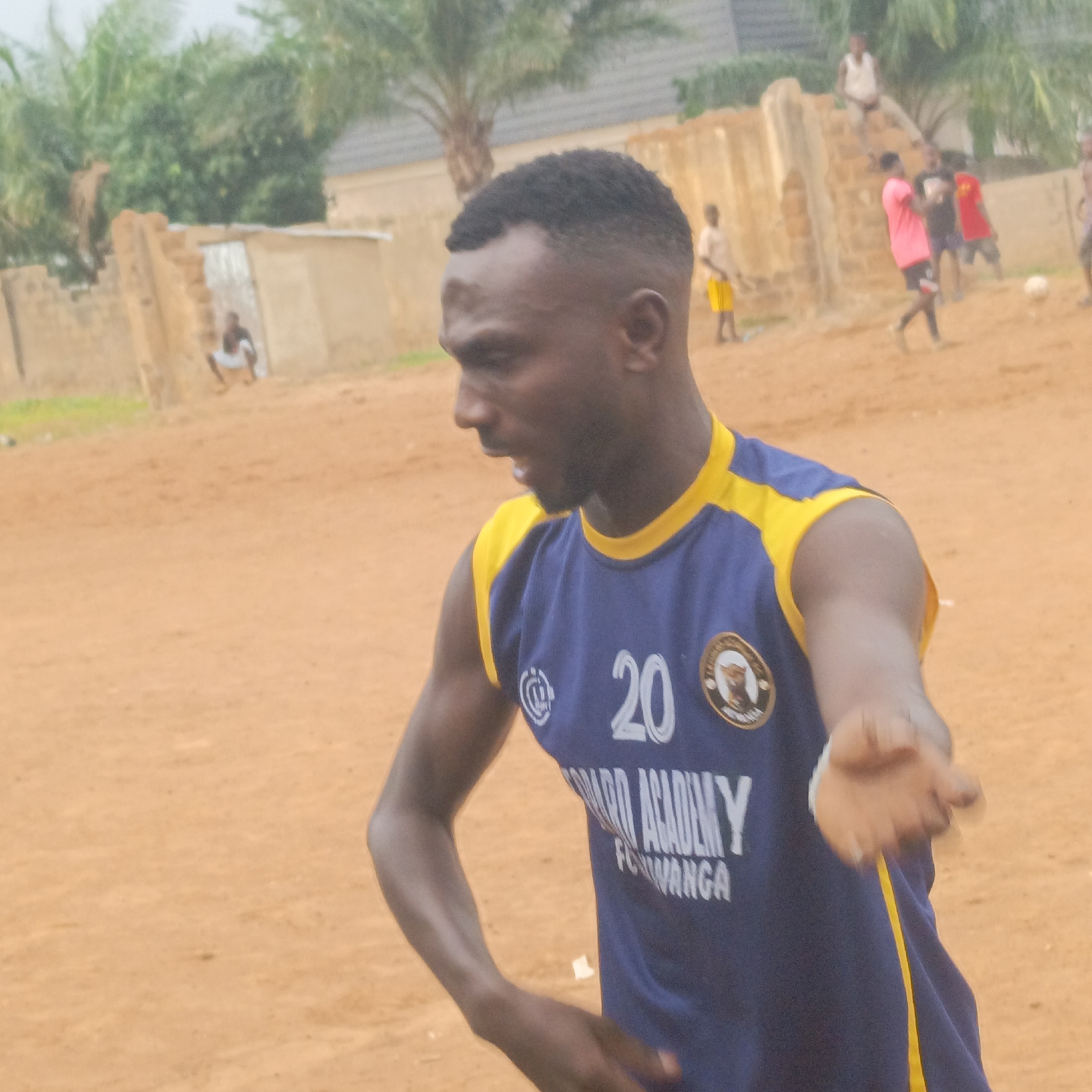 An image captures a young man in a blue and yellow sleeveless jersey, actively engaged in an outdoor setting. He is dark-skinned with a short, neat haircut, looking towards the left side of the frame with his mouth slightly open, suggesting he might be speaking or shouting. His left arm is bent, and his right arm is extended forward, hand open, as if signaling or reaching out. The jersey bears the number "20" prominently in white, along with "WARD ACADEMY" and "AKWANGA" visible below it, indicating a sports team or academy from Akwanga, Nigeria. A circular crest is also visible on the right chest area.
The setting appears to be a reddish-brown dirt or sand field, likely unpaved, suggesting an informal playing area. In the blurred background, several other figures, mostly children or young adults, are scattered across the field. Some appear to be participating in a game, with one figure in a pink shirt and another in a red shirt visible closer to the right. A white football is faintly discernible on the ground to the right of these figures. Further back, simple buildings with low walls and what appears to be a structure made of rough bricks are visible. Palm trees and other foliage can be seen, hinting at a tropical or semi-tropical environment. The lighting suggests daytime, possibly overcast, as there are no harsh shadows. One person is seen squatting near a wall in the far left background. The overall scene conveys a lively, community-based sports activity taking place in a rustic outdoor environment in Akwanga, Nigeria.