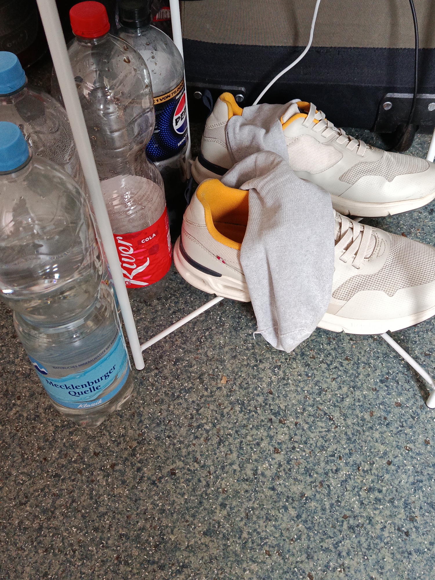 The image shows a close-up of shoes and water bottles. Two light-colored sneakers with yellow accents are in the foreground, with one having a gray sock stuffed into it. Several plastic water bottles and a bottle of cola are visible behind the shoes. The environment appears to be indoors with a speckled floor. The specific location in Halbe, Germany is not ascertainable from the image.