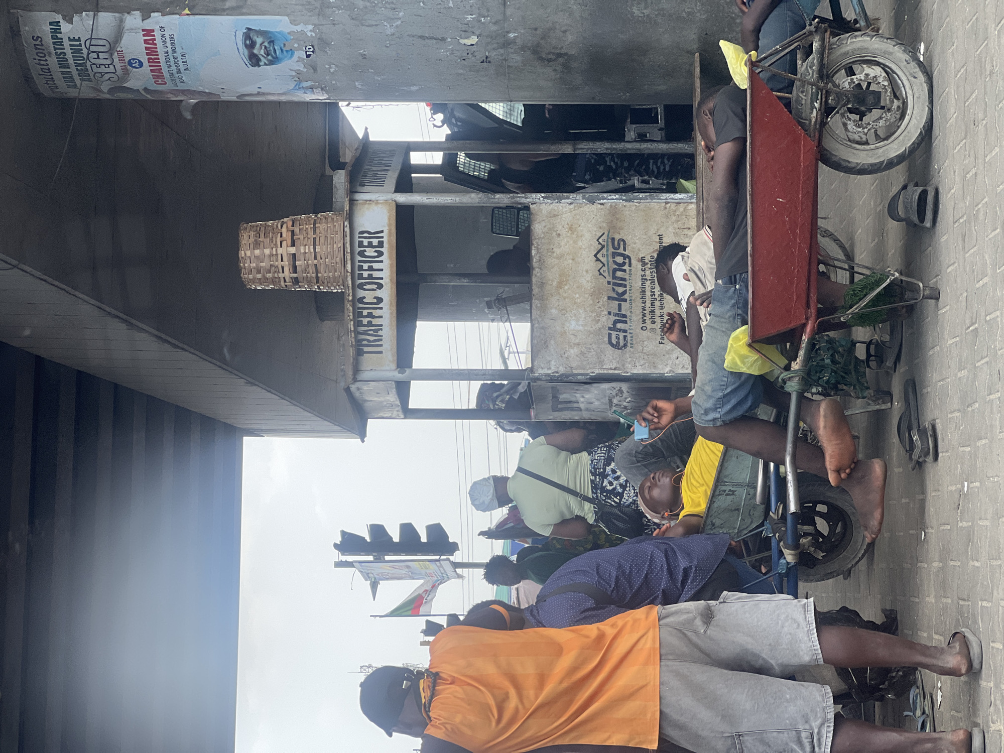The scene depicts a street in Ado, Nigeria, evidenced by the presence of a "TRAFFIC OFFICER" booth with local business advertisement for "Chi-kings Real Estates" and political posters referencing local unions and Chairman elections with the name "ALHAJI MUSTAPHA ABEKUNLE."
Several individuals are present, some lying down and others standing. A three-wheeled handcart is visible with people situated around and on it. One person is wearing an orange shirt and grey shorts. Others are wearing yellow and navy blue shirts. The environment includes a paved street and is partially covered by an elevated structure (likely a bridge). A traffic light is visible.