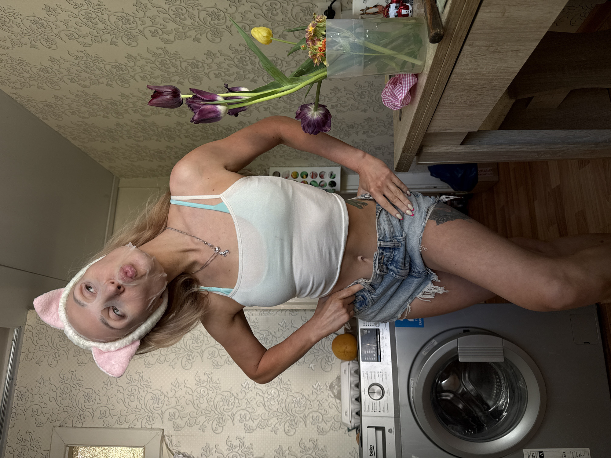 A woman with fair skin, light-colored hair, and wearing a pink cat-ear headband and a white tank top with denim shorts is standing in a room. She has a silver necklace and a light blue bra strap is visible. She has a facial mask on. The background consists of a washing machine, a wooden table with a vase of tulips and other flowers on it, and wallpaper with a floral pattern. The washing machine has a "Beko" logo on it. The photograph was taken in Chaikovsky, Russia.