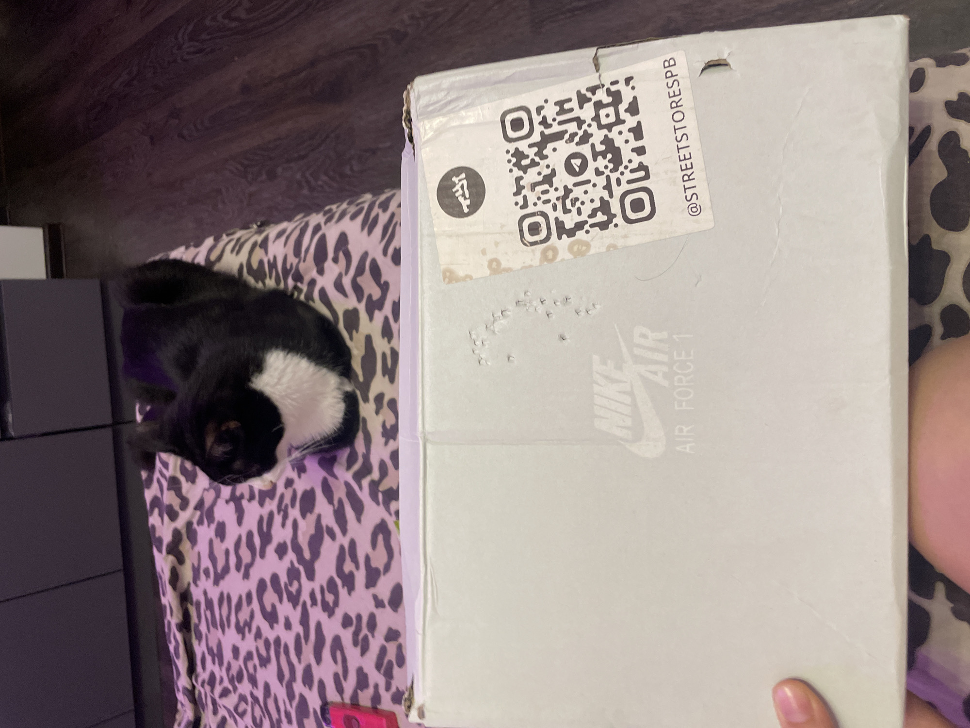 A black and white cat is lying on a bed with a leopard print blanket. In the foreground, a light gray shoebox with "NIKE AIR FORCE 1" printed on it is visible. A QR code and an Instagram handle "@STREETSTORESPB" are on a sticker on the side of the box. The room appears to be a bedroom with wooden flooring and a dark cabinet in the background. The image was taken indoors.