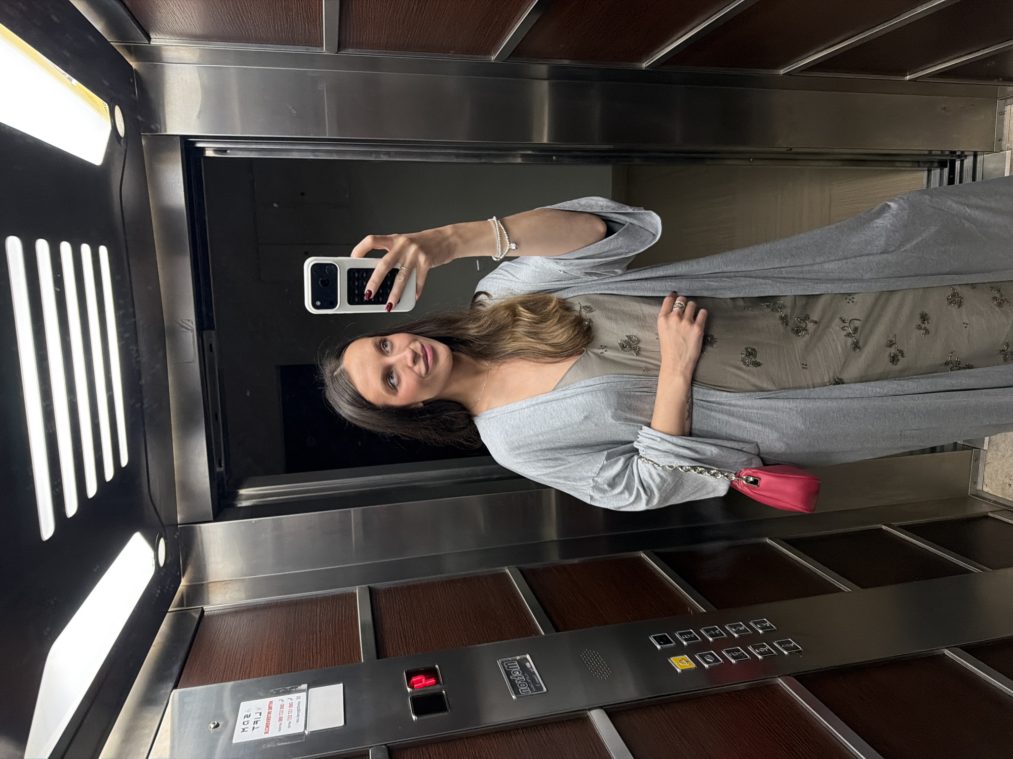 A young woman with long brown hair is taking a selfie in an elevator. She is wearing a light grey outfit and holding a white smartphone with a black border. A small pink purse hangs off her left shoulder. The elevator has a panel with wooden sections and a metal control panel with buttons. The elevator is stopped, as it is reflecting the hallway outside. The specific location in the city of Budva, Montenegro is not discernible.