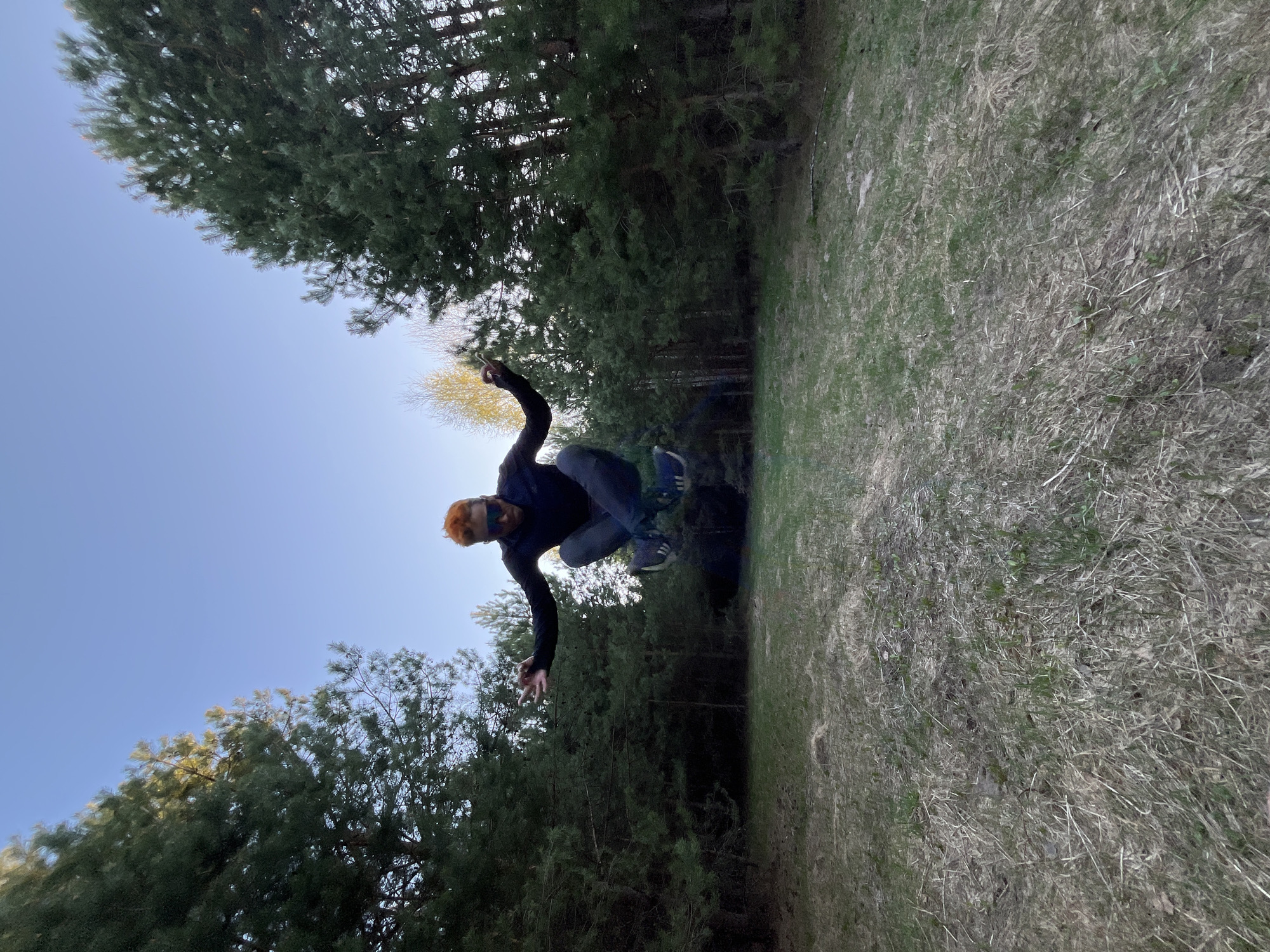 A man with reddish hair is captured mid-air in a forest or park setting, likely in Chaikovsky, Russia. He is wearing dark clothing and appears to be jumping or performing a dynamic movement. The background includes a grassy area and a dense cluster of pine trees. The sky is visible, suggesting it is daytime. The image has been taken from a low angle, emphasizing the height of the jump and the surrounding environment.