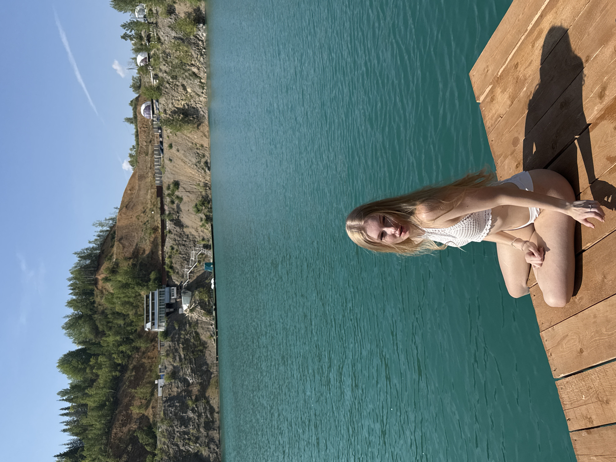 A young, blonde woman is sitting on a wooden dock overlooking a bright turquoise lake. She is wearing a white crocheted bikini top and white bikini bottoms. The lake is surrounded by steep, brown, and green hills. In the distance, on the hillside, there are several buildings and glamping domes. The sky is clear blue with a few wispy clouds. The sun is casting shadows on the dock. The scene is likely located at the Karyer Izvestnyak (Limestone Quarry) in Russia, a popular spot for recreation and tourism due to its striking blue water.