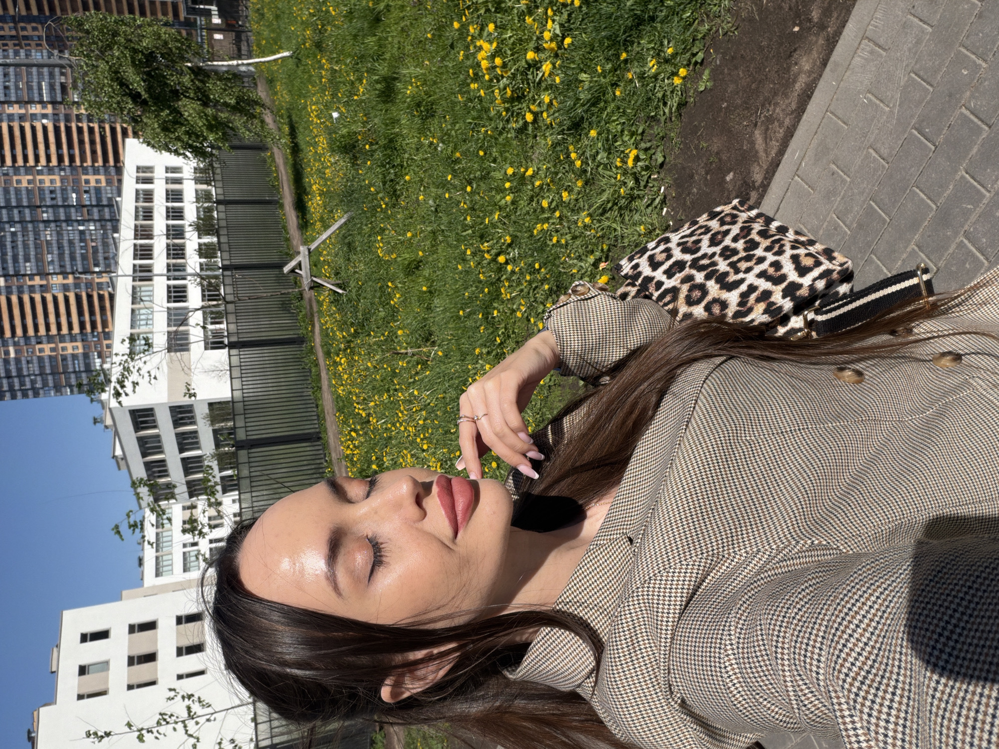 A woman is seen in an eye-level, medium-shot portrait, against an urban backdrop. She appears to be standing near a green lawn with yellow flowers and a building complex. The buildings are characterized by a mix of architectural styles, including modern and possibly Soviet-era designs. The sky is clear and blue. The woman is wearing a brown houndstooth jacket and has a leopard-print bag nearby. Her eyes are closed, and she has long dark hair. She is wearing rings on her fingers. The scene is well-lit, suggesting it is daytime. The location is Unknown, Russia.