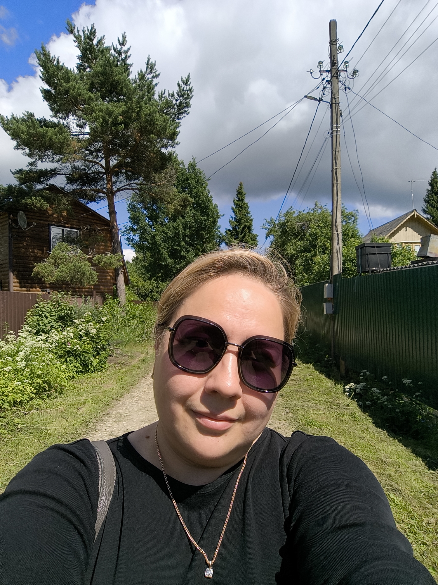 A selfie is taken by a woman with blonde hair, wearing large, dark sunglasses and a black shirt. She is outdoors in a rural setting with a dirt path in front of her. To her left is a wooden house and a brown fence, surrounded by lush green foliage and white wildflowers. To her right, a tall green fence runs along the path. In the background, a utility pole with numerous wires stands prominently against a partly cloudy blue sky. The overall scene suggests a village or countryside location, possibly in Новоникольское, Russia.