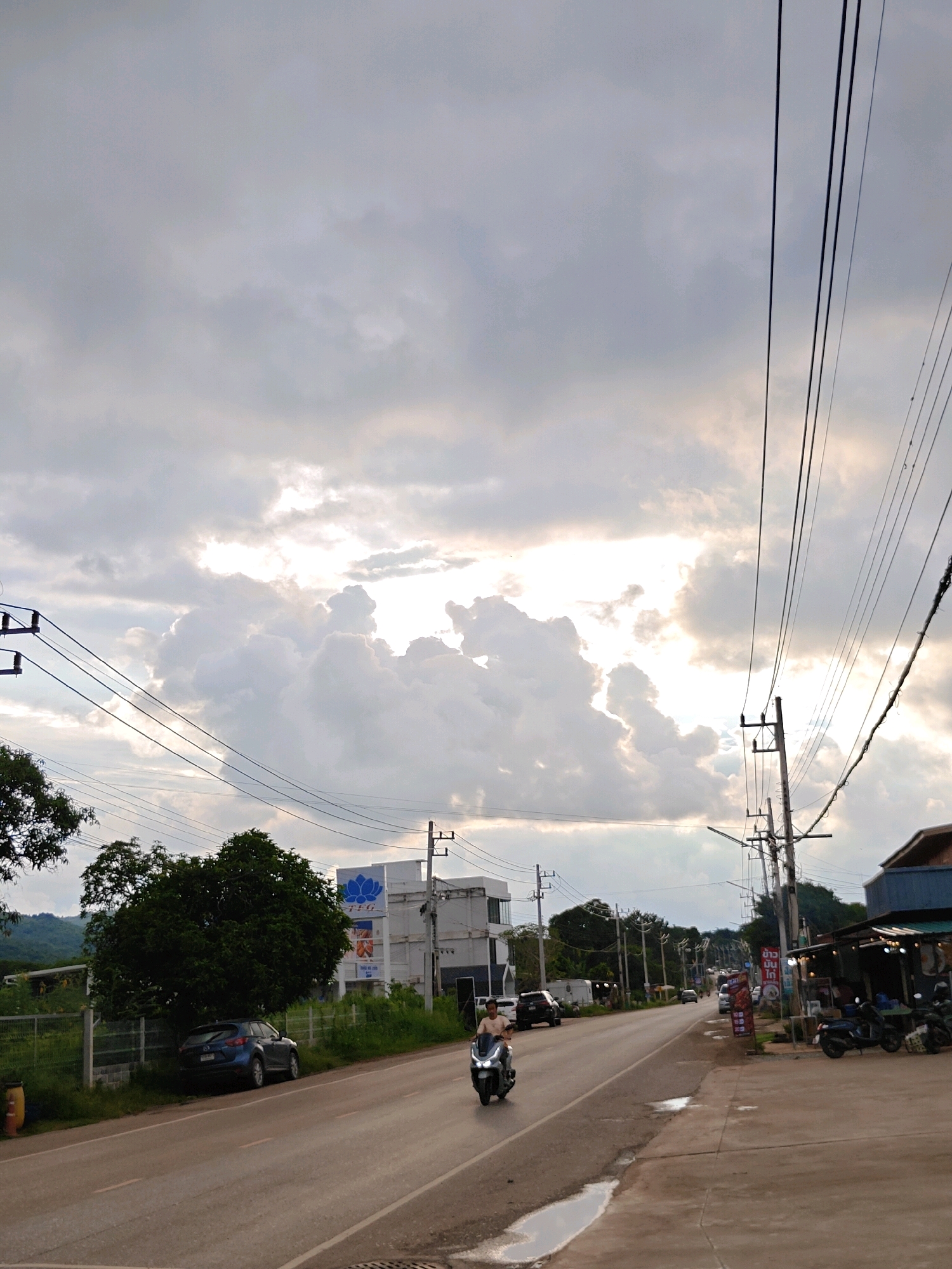 The image depicts a roadside scene in Mu Si, Thailand, under a dramatic sky filled with large, white, cumulus clouds with patches of sunlight breaking through. A two-lane asphalt road runs through the center of the frame, with a concrete sidewalk on the right.
On the left side of the road, a dark blue SUV is parked partially under a large, dense green tree. Further down the road, a white building with a blue lotus logo and the text "TEG" is visible. Numerous utility poles and power lines crisscross the sky.
A person is riding a white scooter in the left lane of the road, moving away from the camera. On the right side of the road, several parked motorcycles are outside a building with a blue roof that appears to be a small shop or restaurant. There are also traffic signs and some vegetation along the roadside. The overall lighting suggests it is either early morning or late afternoon.