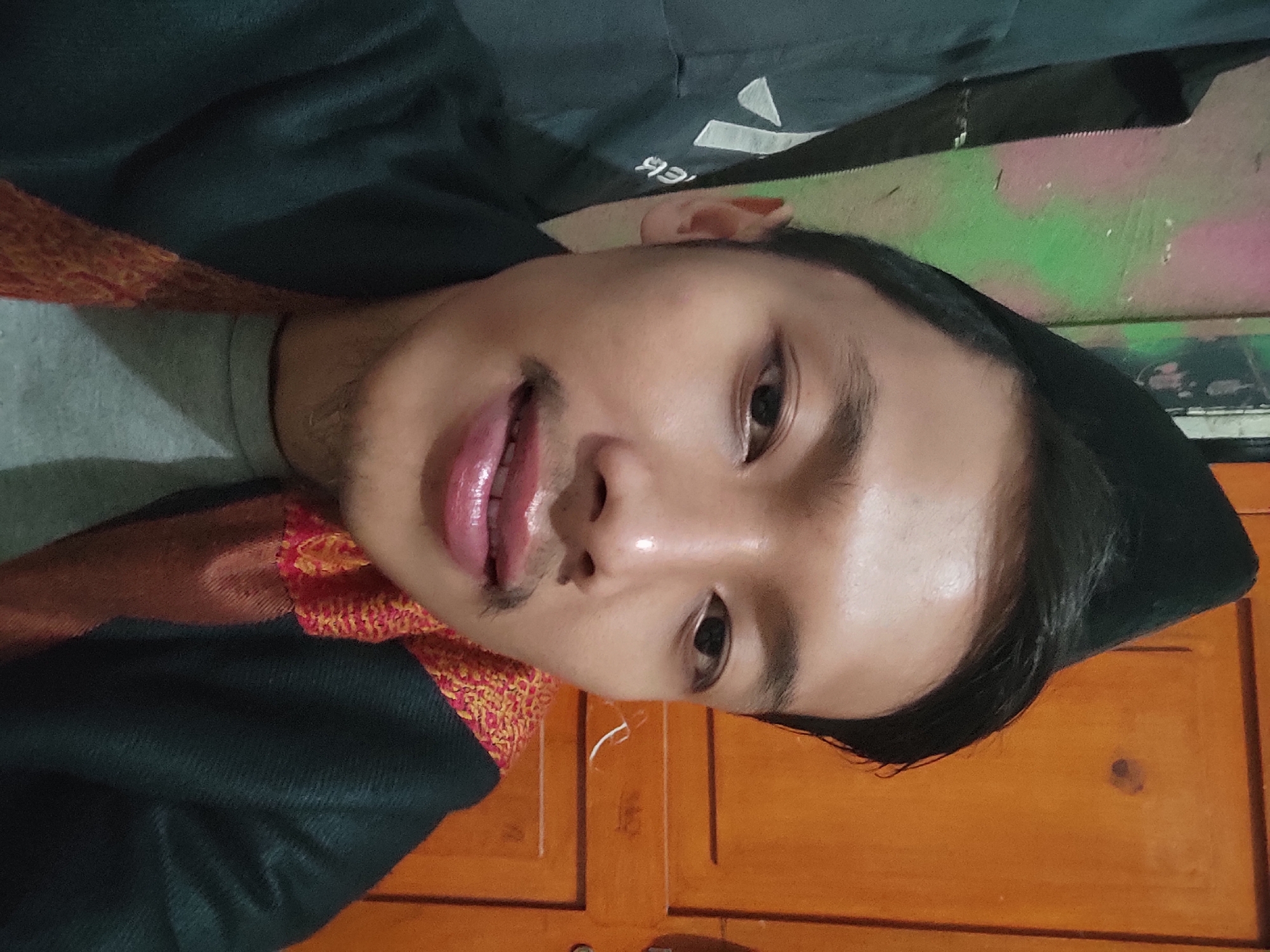 A young man wearing a black peci hat, a grey t-shirt, and a dark shawl with a woven red and yellow trim is pictured. He has a mustache and appears to be looking directly at the camera. The background features a wall with green and pink abstract graffiti and a wooden door. The scene appears to be indoors, possibly in a residence in Sumedang, Indonesia, given the subject's attire and potential cultural context. The lighting is artificial and somewhat dim.
