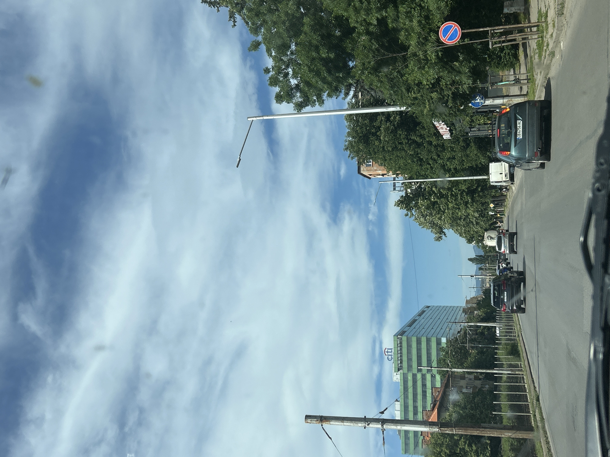 The image depicts a street in Sofia, Bulgaria. The Citi building is visible on the left. Traffic includes several cars and a van. Street signs and utility poles line the road. The sky is blue with scattered clouds.