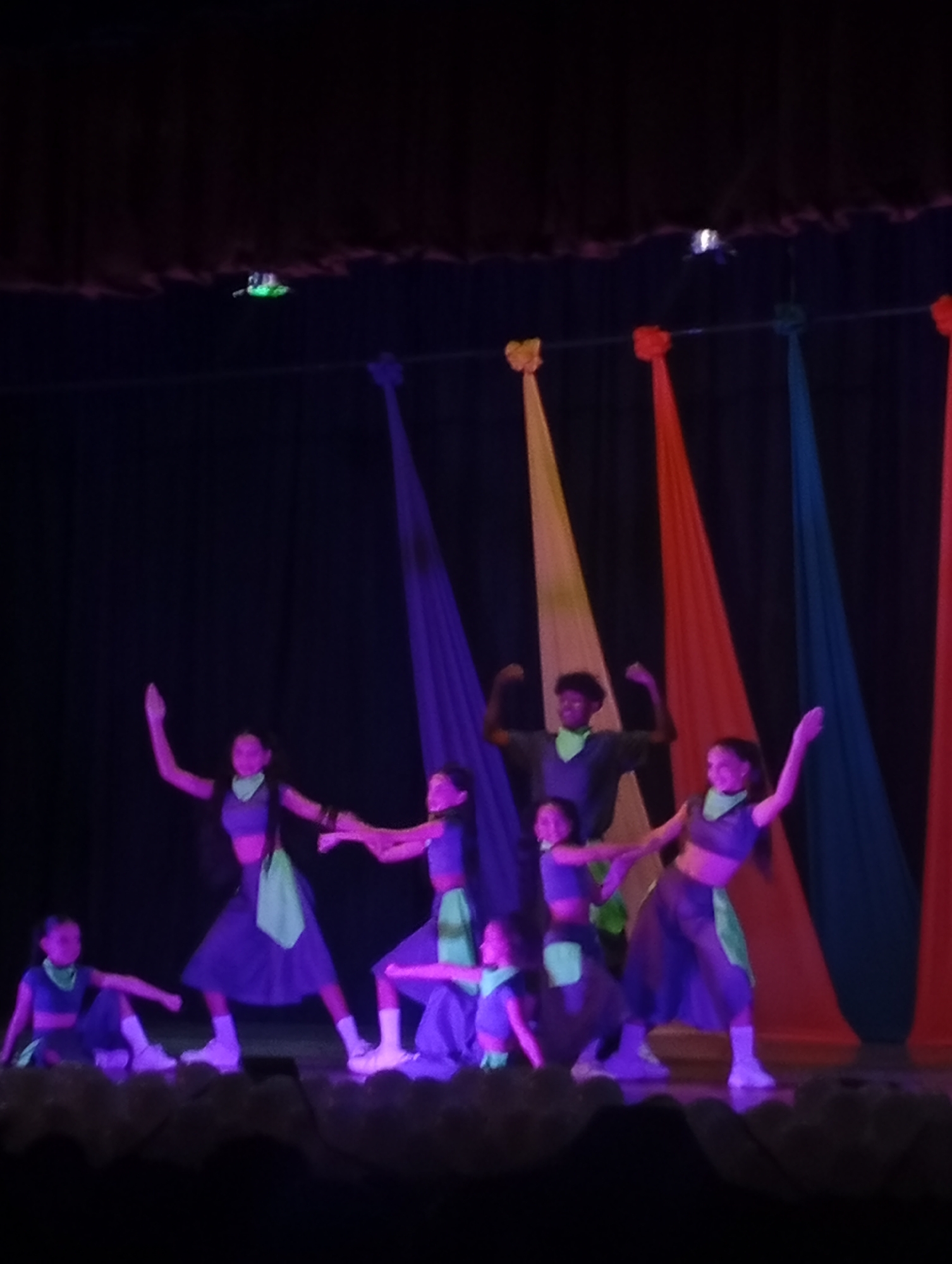 On a stage in Juan Griego, Venezuela, a group of dancers are performing. The dancers consist of both children and adults. They are illuminated by purple stage lights and are wearing coordinated outfits in shades of blue and green with white sneakers. Their performance takes place in front of a black backdrop adorned with colorful fabric panels in yellow, red, and blue. A green light fixture is visible overhead. The scene is dark and the image is of low quality.