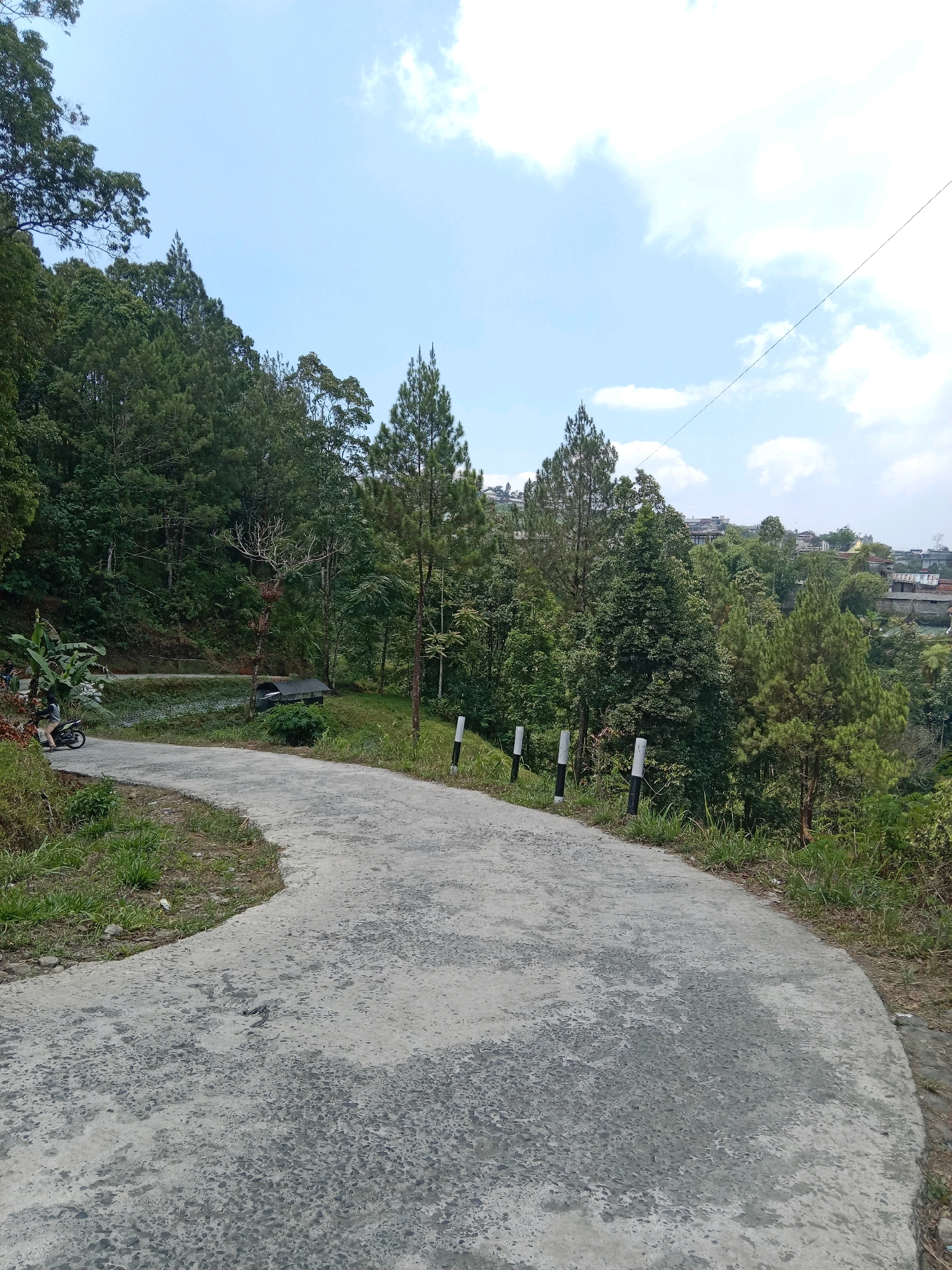 A paved road winds through a lush, forested hillside in Banyumudal, Indonesia. The road is a light gray concrete with visible aggregate. To the left, the hillside is covered in dense green trees, with a few banana plants and a motorcycle parked near the roadside. A person is riding the motorcycle away from the camera, following the curve of the road. Several thin, white posts with black bands are spaced along the right side of the road, marking its edge. Beyond the immediate trees, houses with red roofs are visible nestled on other hillsides, suggesting a populated area within the natural landscape. The sky above is a pale blue with scattered white clouds.