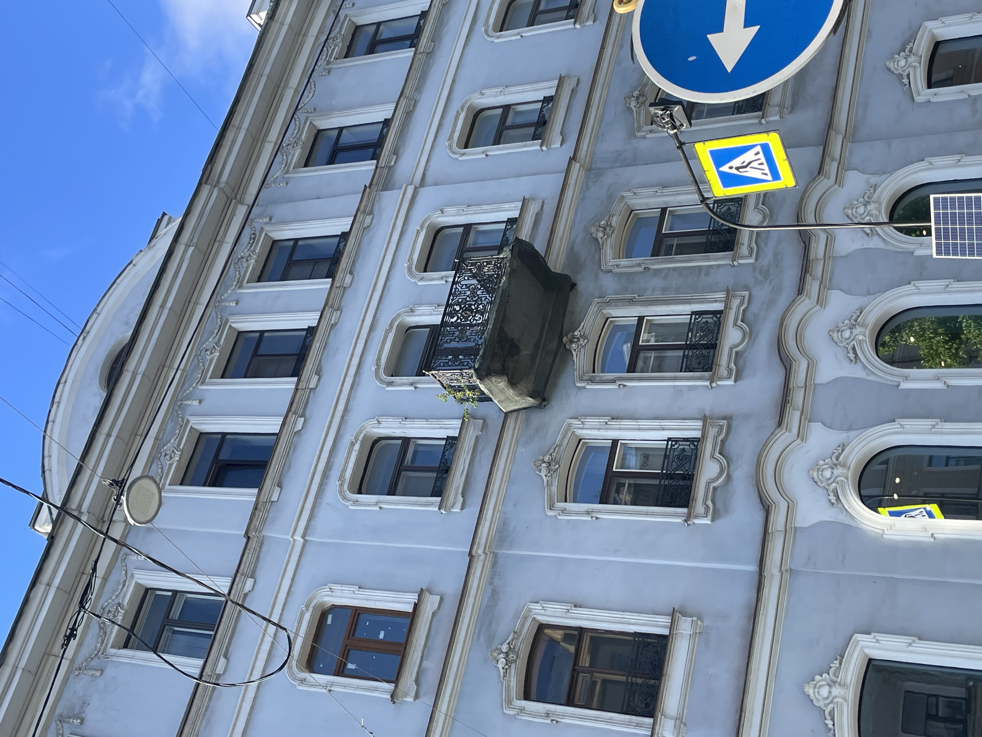 A slightly angled, medium shot reveals the façade of a building in St. Petersburg, Russia. The building, painted in muted tones of blue and gray, features rows of evenly spaced, arched windows framed by decorative white moldings. A single, small balcony with an intricate wrought-iron railing juts out from the building’s surface, adorned with a few small green plants.
Attached to the building, on the right side, are two street signs and a solar panel. The first sign is a blue circle with a white arrow pointing downwards. Below it, there is a yellow square sign with a blue border, displaying an image of a person walking. A small solar panel is mounted beside this sign.
Overhead, a few thin power lines diagonally cut across the top left corner of the frame against a clear, blue sky.