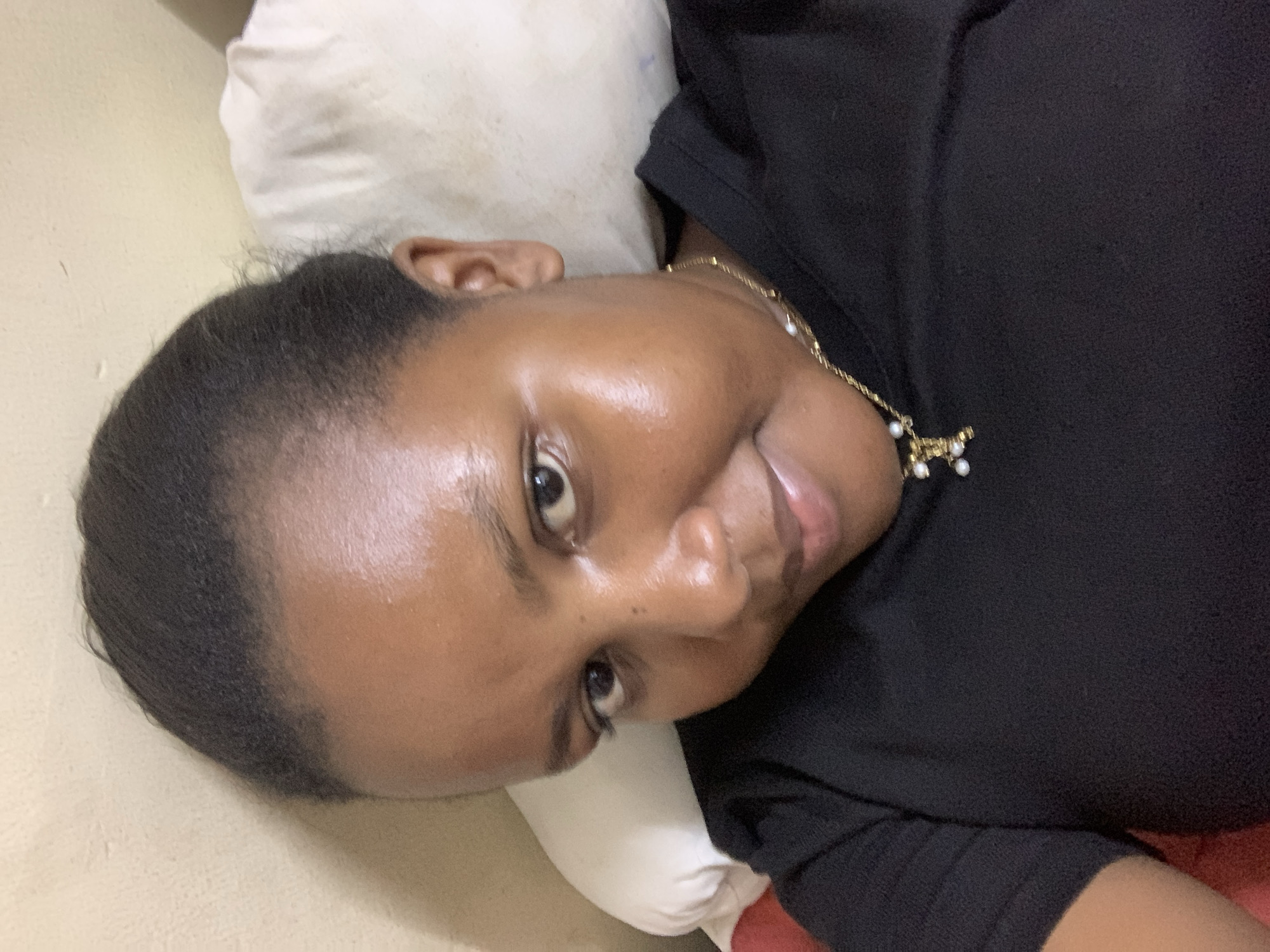 A person lies on a white pillow, wearing a black shirt and a gold necklace with a pearl pendant. Their head is turned slightly towards the camera. The location is unknown.