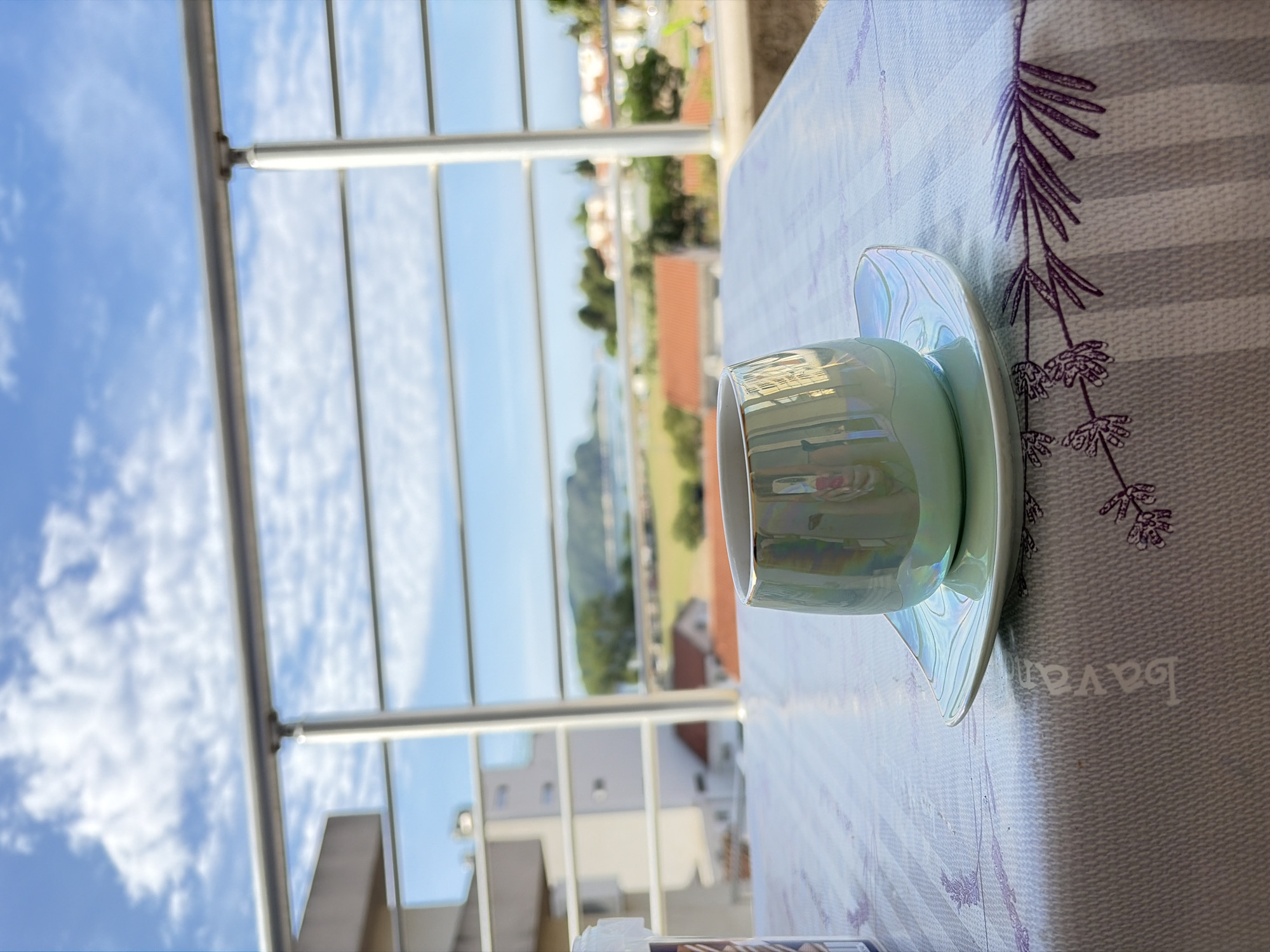 The image shows a light green tea cup and saucer on a striped tablecloth with lavender flower accents. Outside of a fenced balcony, the scene includes a bright blue sky with scattered white clouds, glimpses of buildings, and green vegetation. The location is likely in Budva, Montenegro, given the architecture and vegetation.