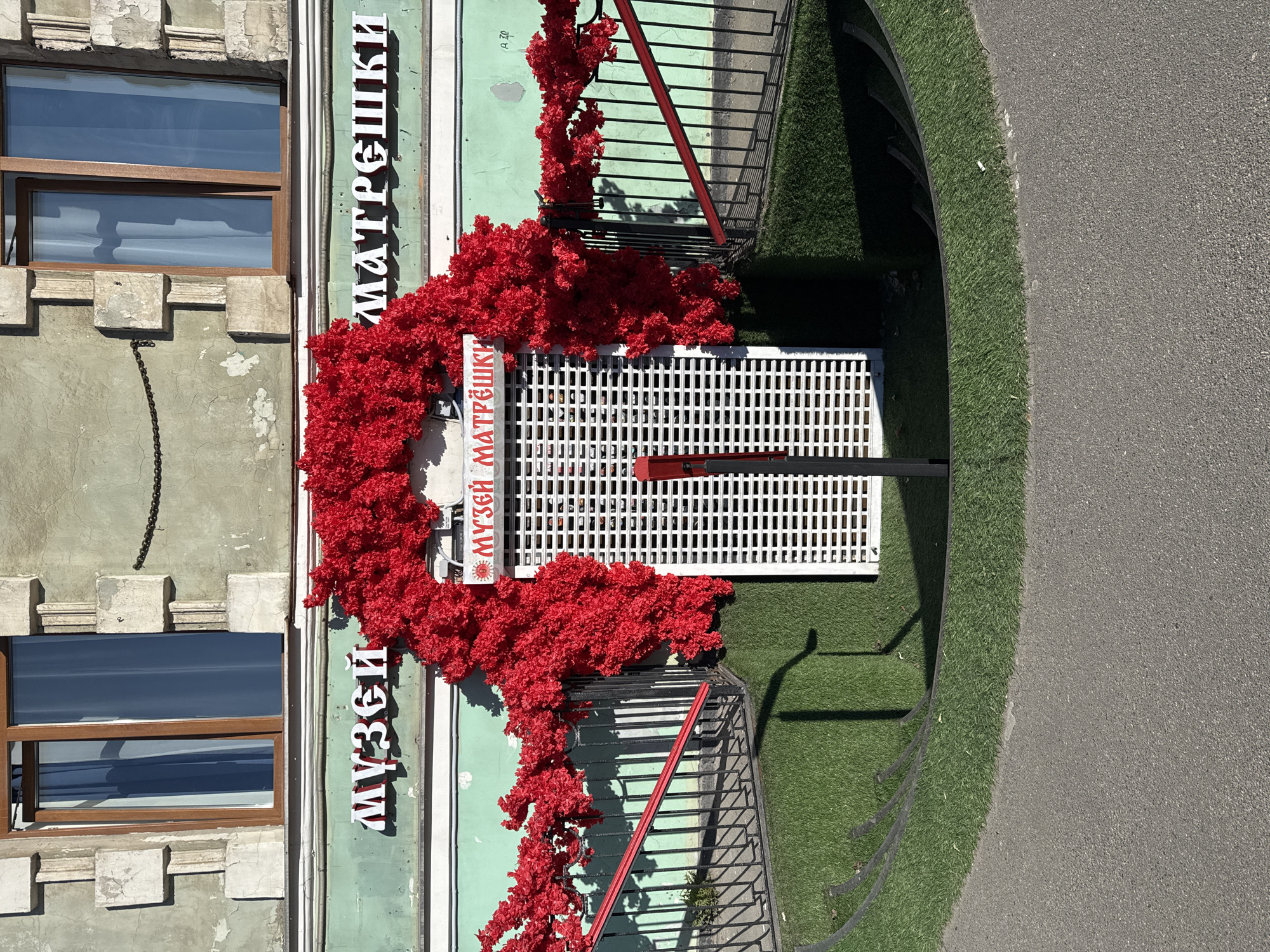 The image depicts the exterior of the "Museum of Matryoshka" in Saint Petersburg, Russia. The facade of the building is light green with vertical white stripes. A large red floral arrangement surrounds the entrance and signage of the museum. The sign displays the name of the museum in Russian. The building has windows with brown frames, and the architectural style appears to be traditional. The ground near the entrance is covered in artificial green turf, adjacent to a paved walkway. A metal railing with red accents is visible, leading to the museum entrance.