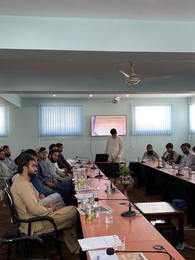 The image captures a scene in what appears to be a conference or meeting room. A group of approximately 15 men are present, primarily seated around a large conference table. The men are dressed in traditional clothing. One man stands at the head of the table near a screen displaying text in what appears to be an Arabic script. A laptop is on the table in front of him. The table is equipped with microphones and littered with documents, water bottles, and glasses. The room has a neutral color scheme with large windows covered by blinds. A ceiling fan is visible overhead. The image is taken in what is believed to be Shurandam, Afghanistan.