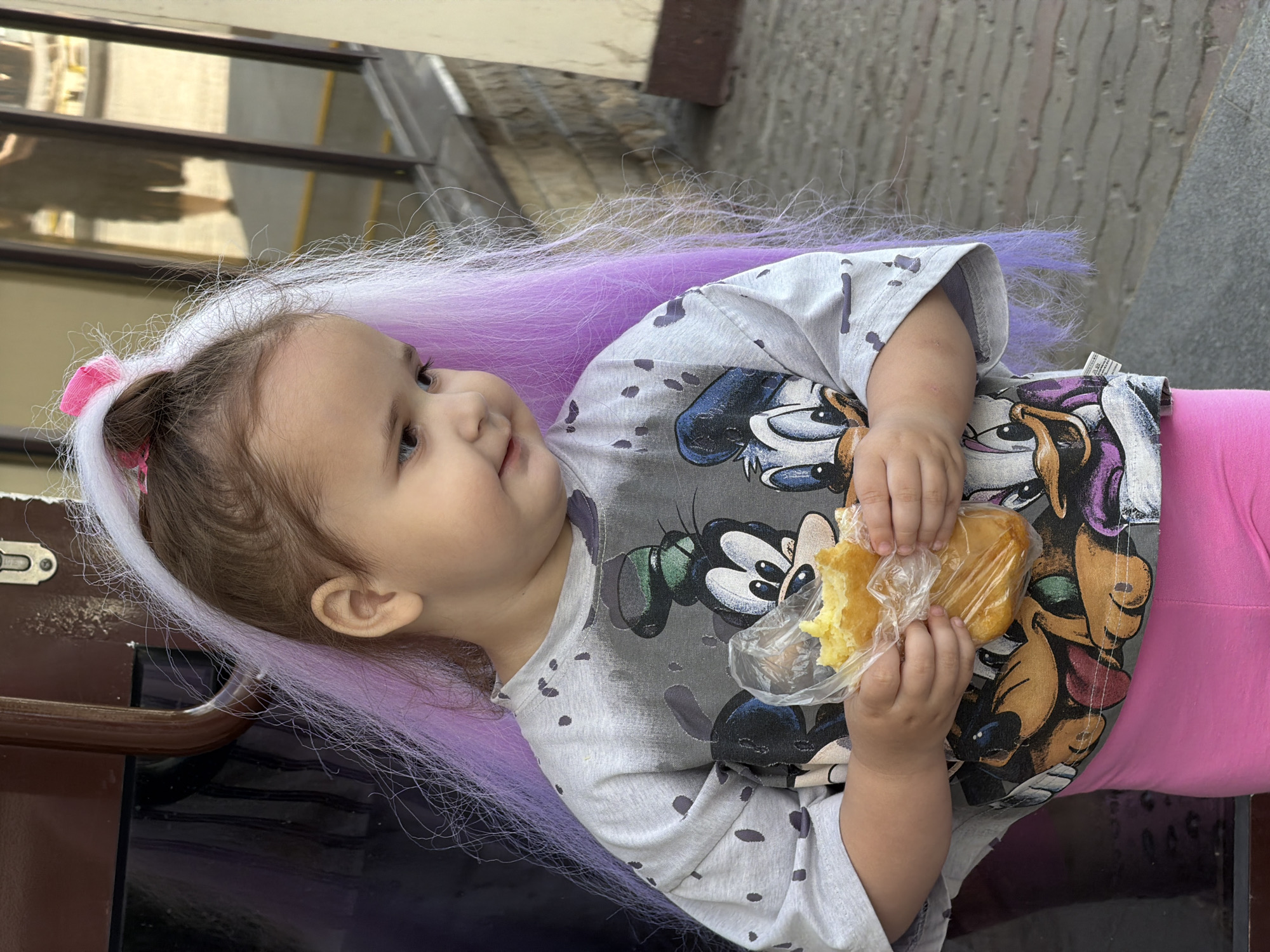 A young girl with a purple wig and a pink bow is standing outside and holding a pastry in a plastic bag. She is wearing a gray t-shirt with cartoon characters on it and pink leggings. The background appears to be a street scene, possibly in Moscow, Russia, with a building and reflections in a window.