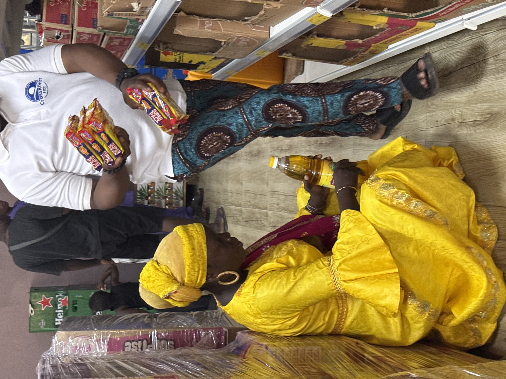 The scene depicts two individuals seemingly shopping at a market in Iranla, Nigeria.
One person is wearing a white t-shirt with the word "CrossRiver", printed pants, and sandals, and is holding several packs of biscuits. The other person is wearing a yellow traditional garment and headwrap, and holds a bottle of what appears to be cooking oil. They are leaning atop a wrapped pallet of retail goods, which sit on what looks to be a pallet of beer crates. Shelves filled with boxed goods are visible in the background.