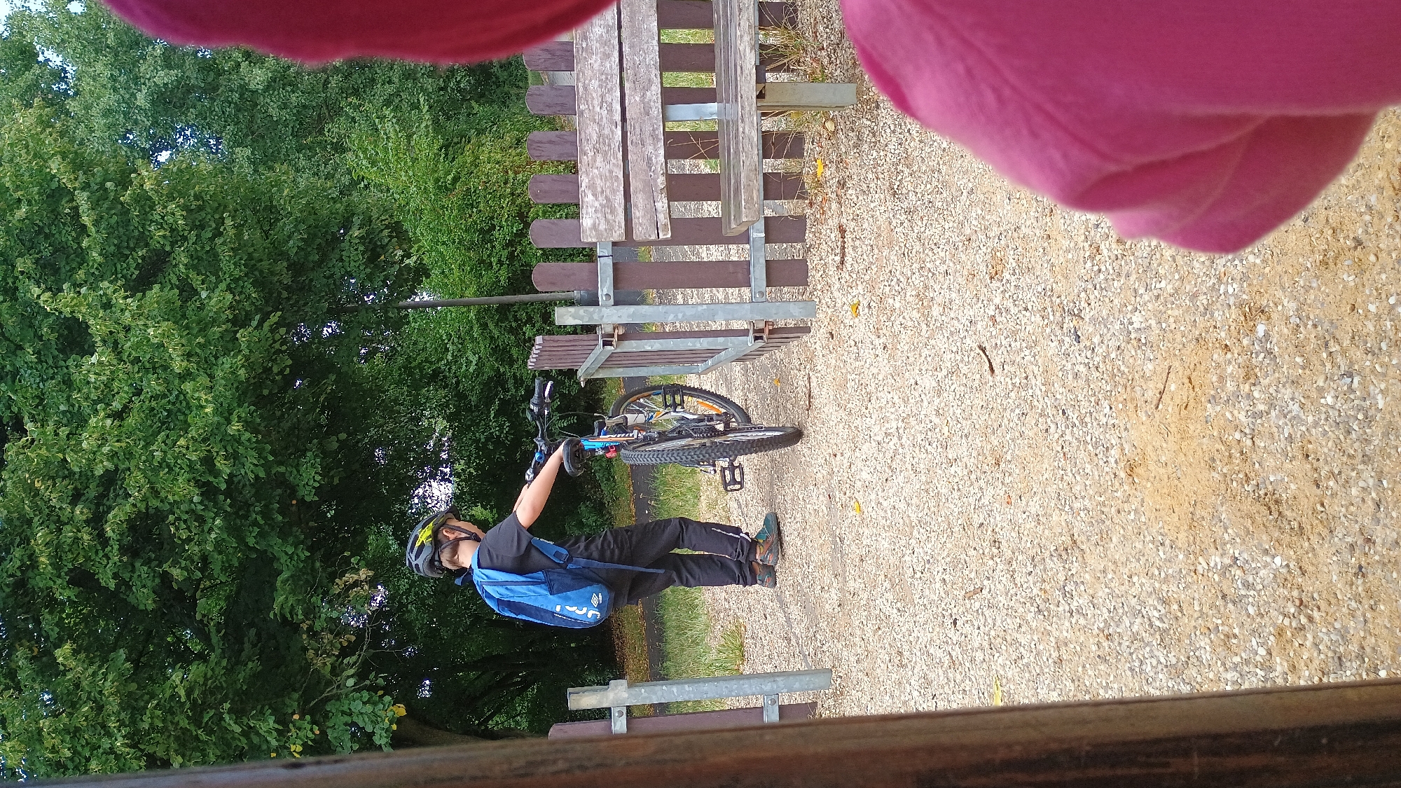 A young boy wearing a blue backpack and a helmet is standing next to a bicycle in a park-like setting. He is dressed in black pants and a black t-shirt. The bicycle is blue and black with orange accents on the spokes. The boy is holding the handlebars and appears to be either preparing to ride or has just stopped. The ground is covered in gravel. In the background, there is a wooden fence and a wooden bench. Behind the fence are lush green trees. The scene is outdoors, likely on a path or trail, with the gravel suggesting an area that experiences a lot of foot and bike traffic. There is also a pink garment visible in the foreground, which appears to be part of another person's clothing, suggesting the image was taken by someone else who is close by. The setting is consistent with an outdoor recreational area in the city of Sokolov, Czechia, possibly a park or a cycling path.