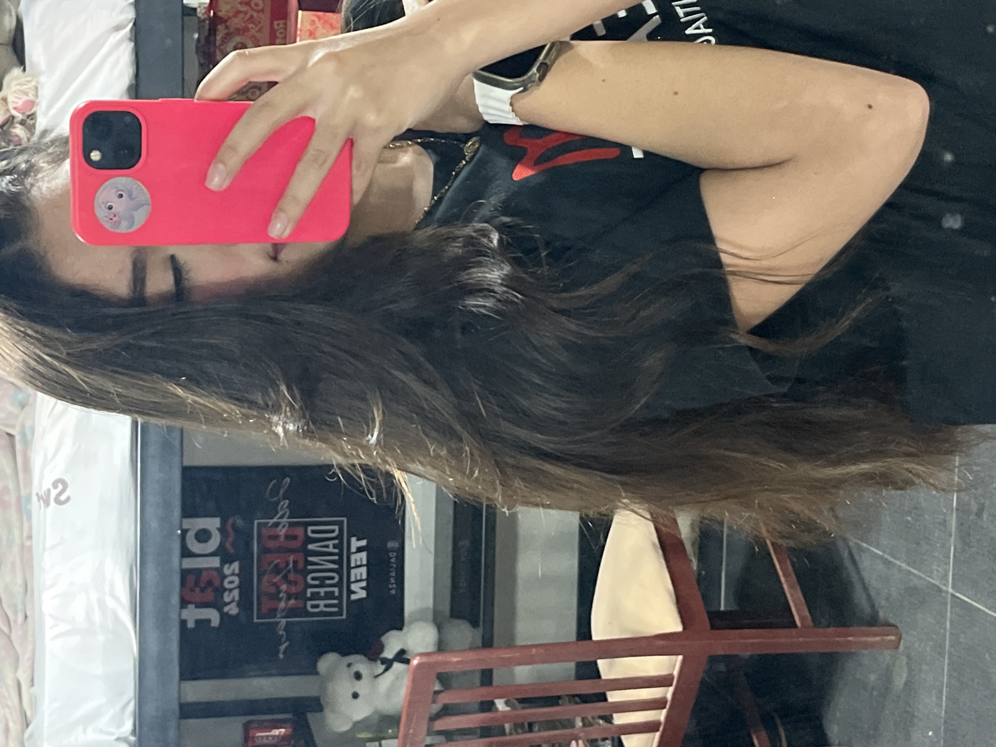 A young person is taking a mirror selfie in a room. They are holding a bright pink smartphone in their right hand. The phone has a circular sticker on the back. The person has long, dark brown hair, which partially obscures their face. They are wearing a black t-shirt with a red graphic design and a necklace. A white watch is visible on their left wrist.
In the background, a red chair with a cream-colored cushion is visible. Behind the chair, a dark banner is hanging on the wall. The banner has text that is partially obscured and appears to be in a foreign language. A white teddy bear is also visible on a shelf to the left of the banner. Above the banner, a white mattress or bedding is seen. The floor appears to be tiled in a dark color. The scene suggests an indoor residential setting.
The location is identified as Juan Griego, Venezuela.