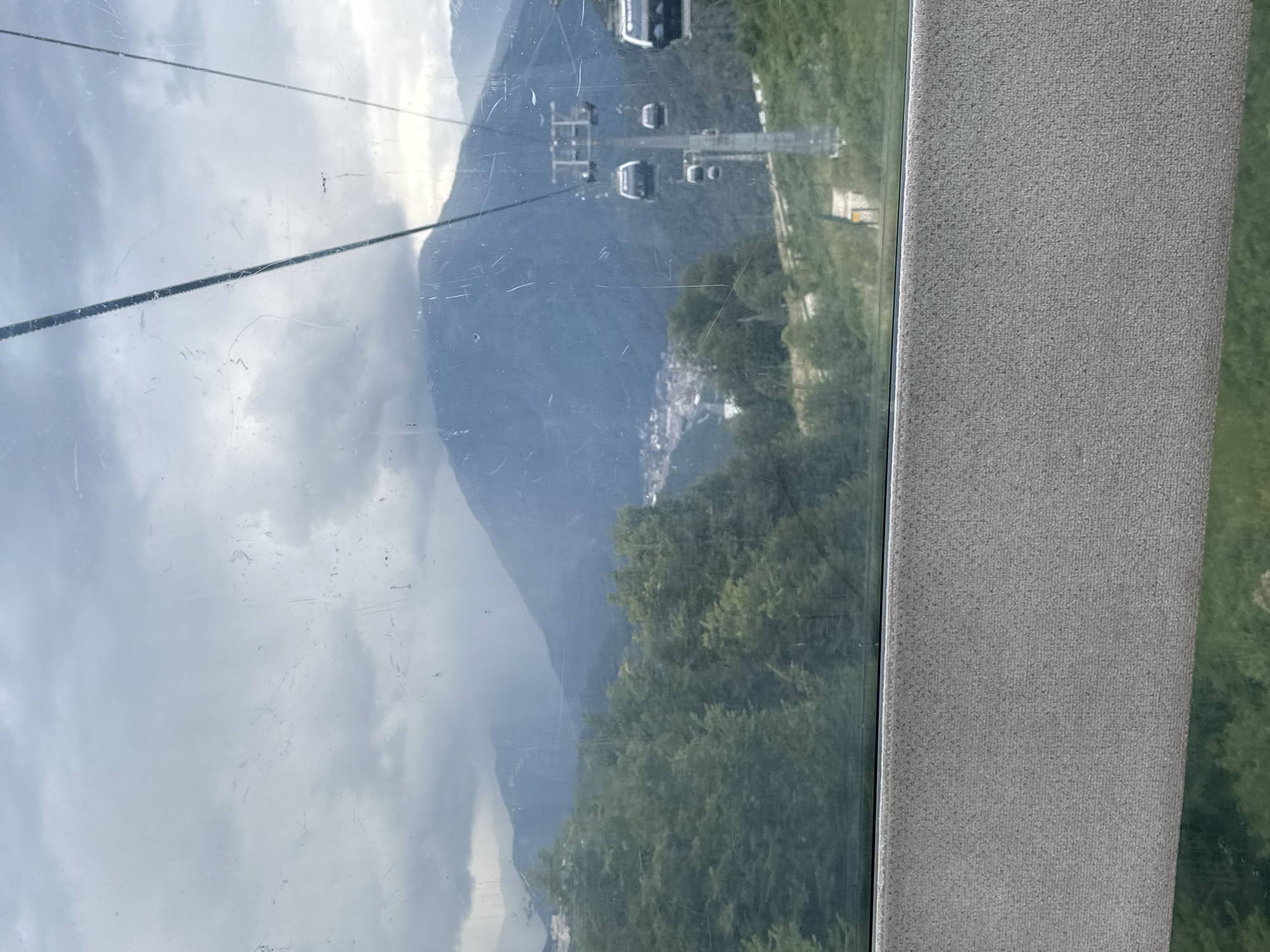 The image shows a view from a cable car overlooking the mountain resort town of Estosadok, Russia. The cable car cabins are visible in the foreground and midground, suspended from wires. Below, a dense forest of green trees covers the lower slopes of a large, misty mountain. In the distance, a small town with buildings can be seen nestled in a valley. The sky is overcast with heavy clouds. The view suggests a cool, possibly rainy day. The foreground of the image includes a grey textured surface, likely the interior of the cable car.