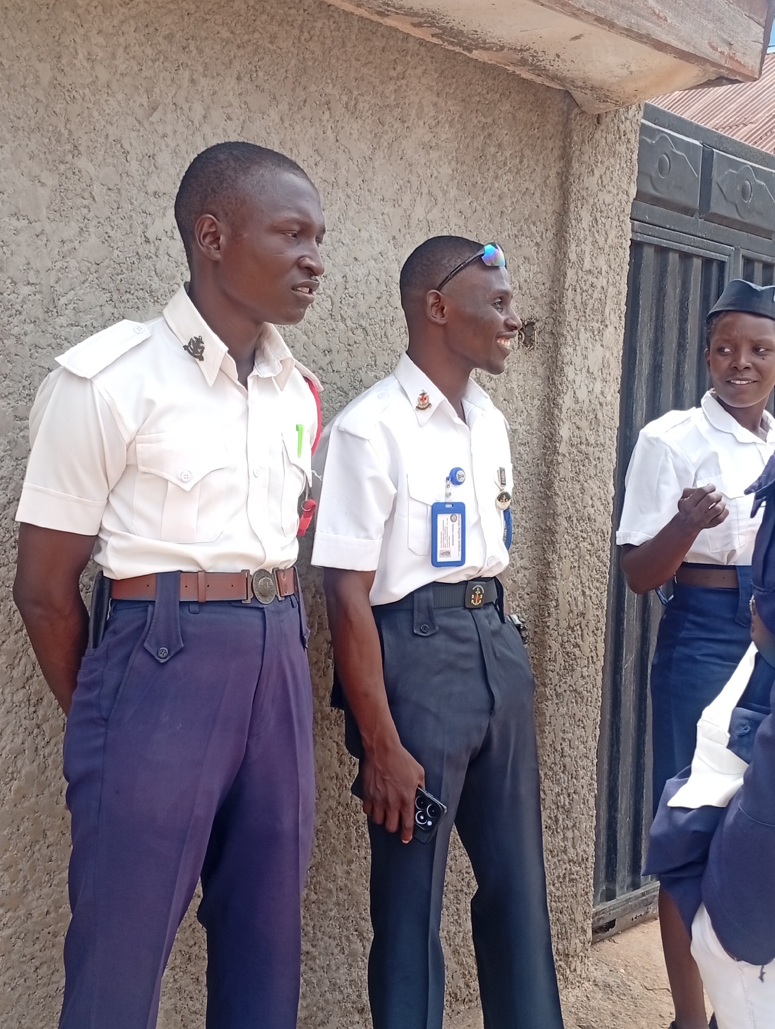 The image shows three individuals standing outdoors. Two men in matching uniforms consisting of short-sleeved white shirts and dark pants with prominent belts are in the foreground. The man on the left has his hands clasped behind his back and a brown belt. The man on the right is holding a smartphone and wearing sunglasses on top of his head with a dark belt. A woman in a similar uniform, including a small dark hat, is partially visible in the background near a dark metal gate. The location appears to be outdoors, with a textured beige wall behind the men. The city and specific location are unknown.