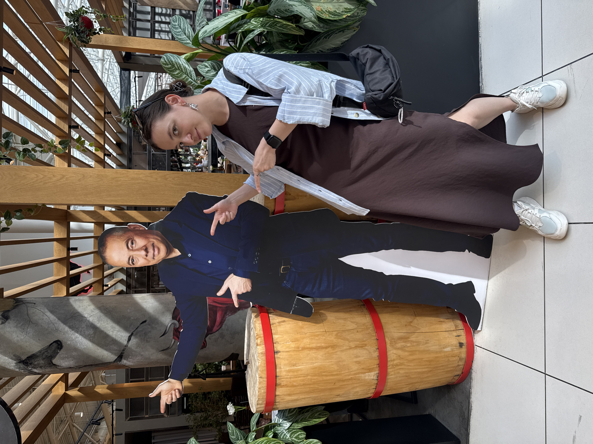 The photo shows a woman posing with a cardboard cutout of a man in what appears to be an indoor public space, likely a shopping mall in Khimki, Russia. The woman has dark hair and is wearing a brown dress with a light blue striped shirt over it. She has a black bag slung over her shoulder and is wearing white sneakers. She is pointing toward the cardboard cutout with both hands. The cardboard cutout, positioned on top of a wooden barrel, depicts a man in a blue shirt and jeans, pointing with both hands. The environment includes indoor plants, wooden beams, and what seems to be the interior architecture of a mall, possibly in the city of Khimki, Russia.