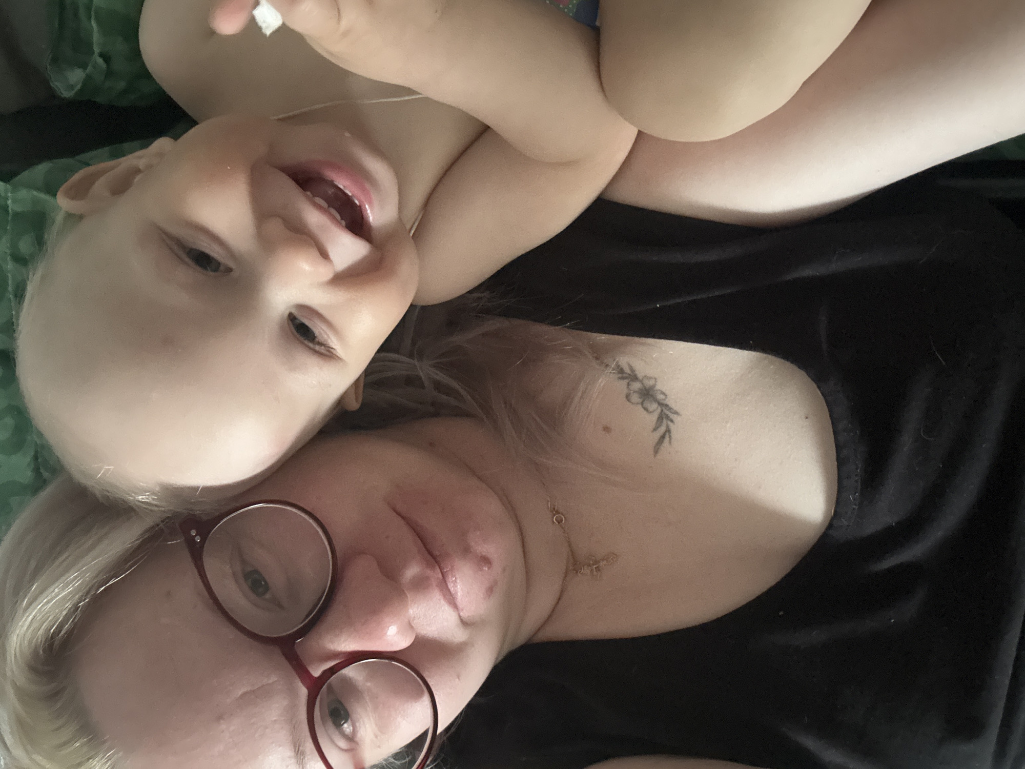 An intimate, close-up portrait shows an adult and a baby/toddler lying down, their faces captured from an overhead, slightly rotated perspective. The scene likely takes place indoors in Chaikovsky, Russia. In the foreground, on the left side of the frame, is an adult with blonde hair and red-framed glasses, their eyes looking towards the camera. They wear a black top, and a small floral tattoo is visible on their upper chest, along with a thin gold necklace. Their expression is gentle and content. Above and to the right, leaning on the adult, is a happy baby/toddler with light hair, smiling widely with an open mouth, showing their teeth. One of the baby's hands is near their face, holding a small white object, possibly a piece of tissue or paper. A green patterned fabric, likely a blanket or pillow, is visible in the background on the far left. The image evokes a warm and affectionate bond between the two subjects. No visible text is present in the image.
