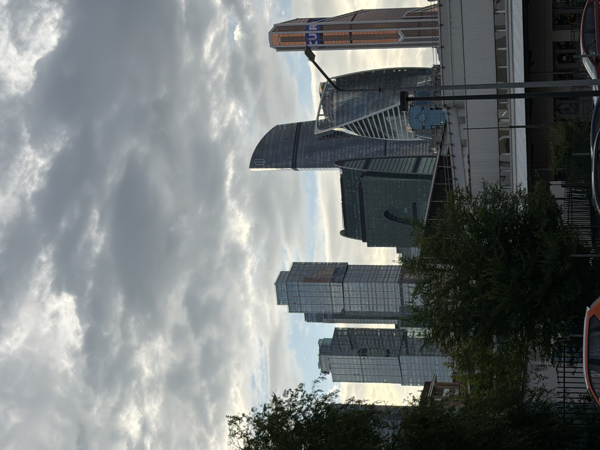 This is a photograph of the Moscow International Business Center, also known as Moscow City, a commercial district in the western part of Moscow, Russia. The image showcases a cluster of modern skyscrapers with distinctive architectural designs. The sky is filled with dramatic, overcast clouds, suggesting either early morning or late afternoon light.
In the foreground, the tops of trees and foliage from a park or green space are visible on the left, partially obscuring the lower portions of some of the buildings. Below the skyscrapers, there is a multi-story structure with a white facade and a metallic roof, possibly a transportation hub or a commercial building. A street lamp is positioned in front of some of the buildings.
Several cars are partially visible at the bottom of the frame, suggesting this is a street-level view looking up at the city skyline. The cars are predominantly modern passenger vehicles. The overall atmosphere is one of an urban landscape under a moody sky. No people are discernible in the image. The scene is static, capturing a moment in time without any discernible action.