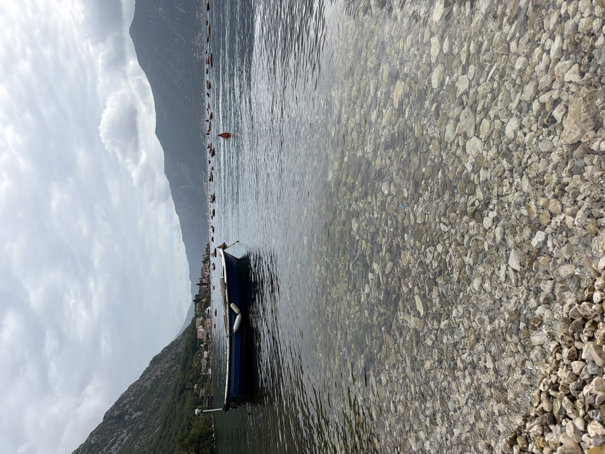 The image captures a serene view of the Bay of Kotor in Donji Orahovac, Montenegro. The foreground showcases a rocky shoreline merging into clear water, revealing the seabed's stones. A small blue boat is moored in the water, close to the shore. A row of red buoys stretches across the water in the mid-ground. In the background, a towering mountain slopes towards the bay, and a stretch of buildings. The sky is overcast.
