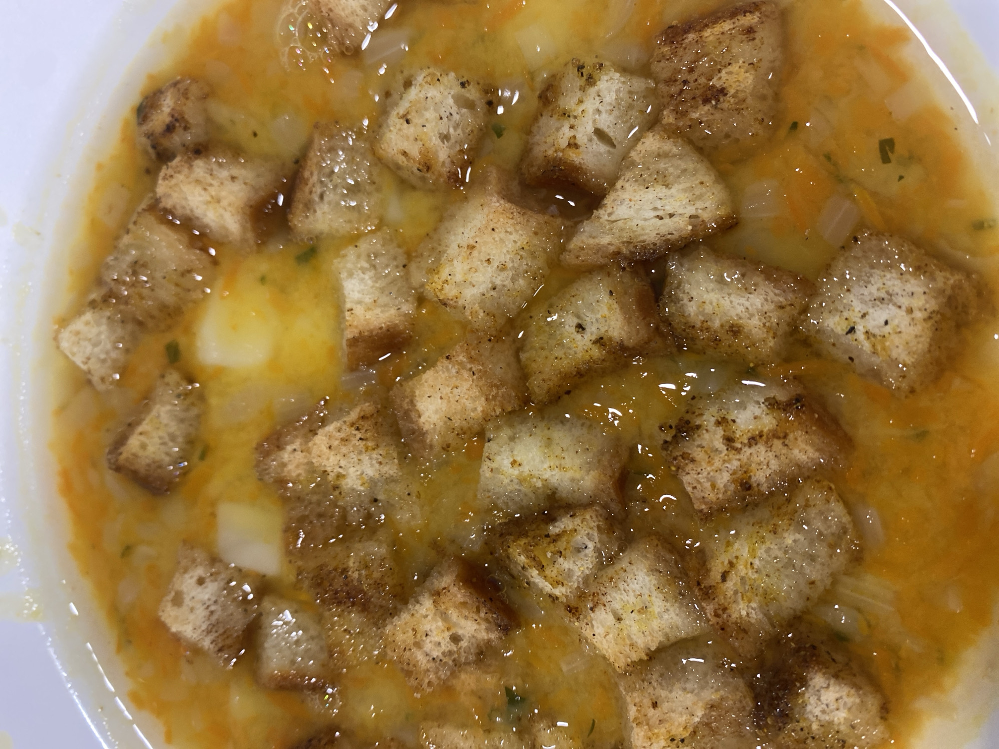 The image is a close-up, overhead shot of a bowl of soup. The soup is a bright orange color and appears to be chunky with pieces of what look like potatoes and carrots. It is topped with numerous toasted croutons. The lighting suggests indoor conditions, possibly a restaurant or home kitchen. There are no people visible in the image, and no specific geographical markers indicate it is located in Rozhny, Ukraine.