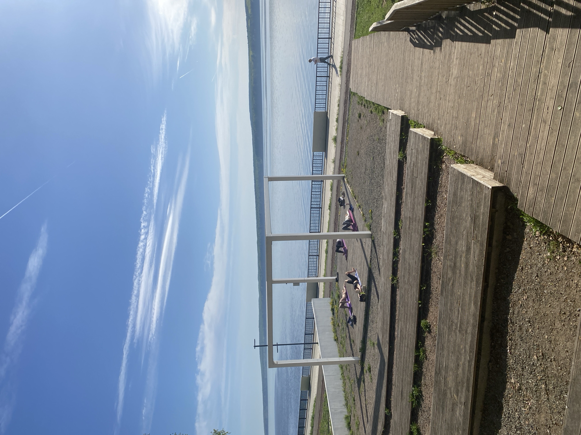 Captured from an elevated vantage point, the photograph presents a scene in Chaikovsky, Russia, near a waterfront. The environment includes a body of water, likely a river or a large lake, under a clear blue sky marked with contrails. A wooden boardwalk or path extends along the right side of the frame, with wooden steps descending towards a lower level where a series of white, rectangular structures stand.
Several individuals are engaged in what appears to be a yoga or exercise session on the ground near the white structures. They are positioned on mats, suggesting preparation for physical activity. Further along the waterfront promenade, a lone figure is walking, seemingly enjoying the view.
The immediate surroundings include patches of gravel and greenery interspersed with the wooden walkways, hinting at a park-like or recreational area. A metal railing runs along the edge of the waterfront, providing a safety barrier.