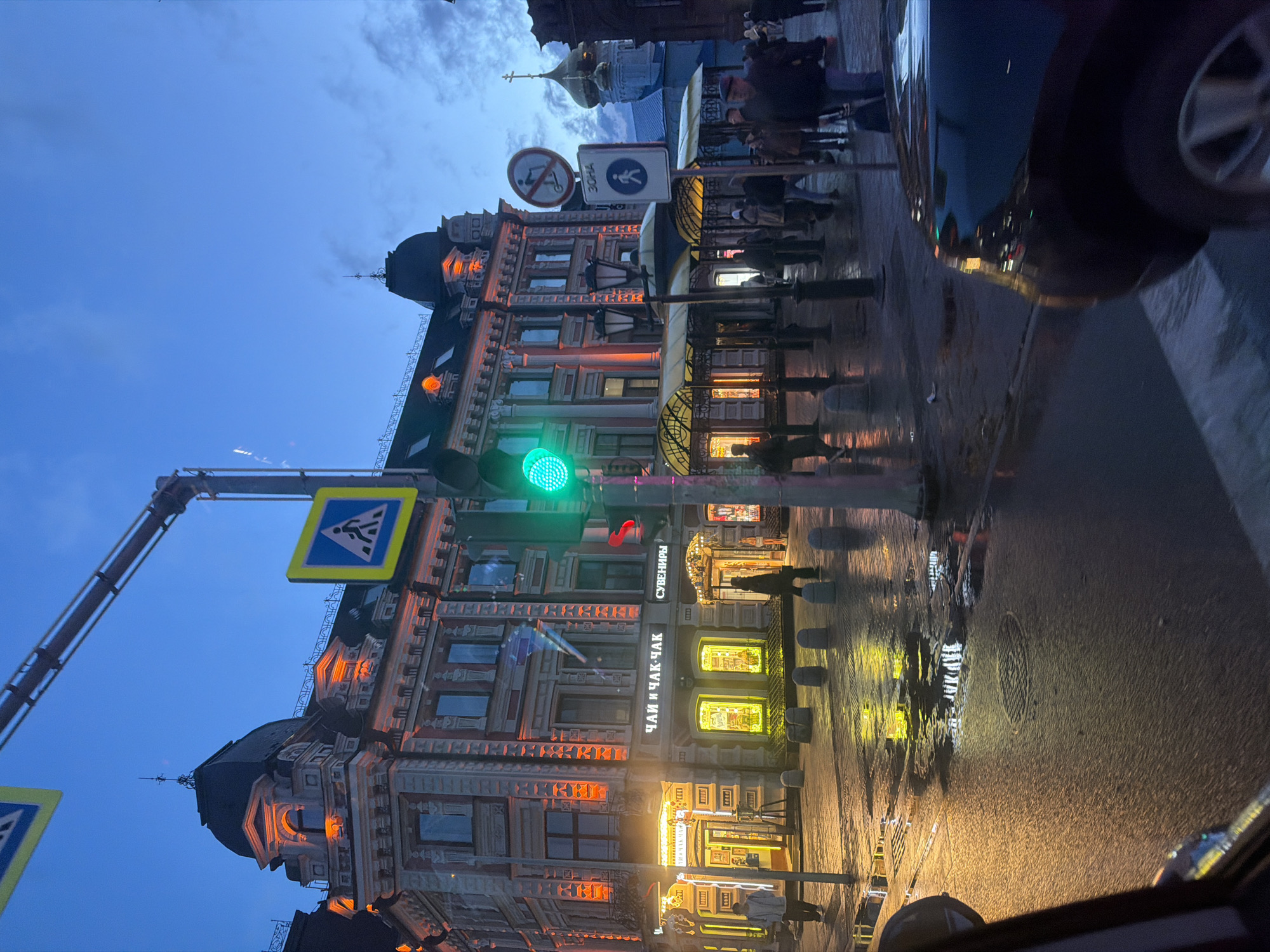 This image captures a vibrant street scene in Kazan, Russia, at dusk or night, under wet conditions. The view is from a low perspective, possibly from inside a vehicle, with part of a car visible on the right edge.
In the midground, a grand, multi-story building with ornate architecture dominates the left side. Its windows emit a warm, orange-yellow glow, reflecting on the damp sidewalk. Visible signs on this building include "ЧАЙ И ЧАК-ЧАК" (Tea and Chak-Chak), indicating a shop selling traditional Tatar sweets, and "СУВЕНИРЫ" (Souvenirs). A brightly lit green traffic light is prominent in front of the building, alongside two blue pedestrian crossing signs. Further down the street, people are visible walking along the sidewalk, some as silhouettes, others partially illuminated by the ambient light.
The foreground features a wet asphalt road, reflecting the artificial lights and parts of the surrounding buildings and vehicles, with a visible manhole cover. Along the sidewalk, several light-colored bollards are evenly spaced.
In the background, under an overcast blue-grey sky, a golden dome topped with a cross, likely belonging to a church or cathedral, is discernible above the buildings. Additional street signs are present, including a red-circled sign prohibiting scooters, with the word "ЗОНА" (Zone) next to it.
The damp surfaces create striking reflections, enhancing the urban atmosphere of this busy street. The overall impression is an active city environment lit by artificial lights against the twilight sky.