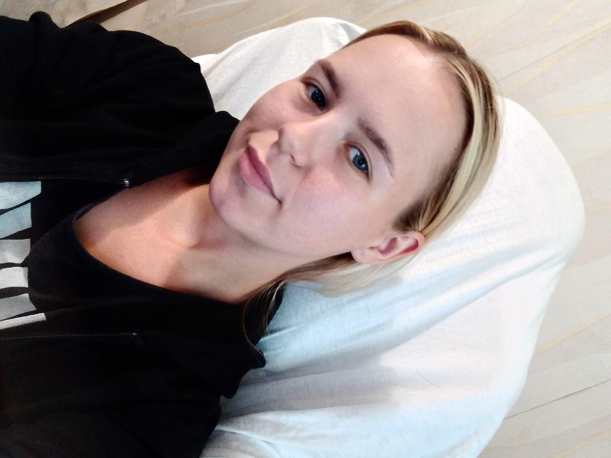 This is a medium shot, selfie-style photograph of a young Caucasian woman lying down on a white pillow and bed. She has fair skin, blonde hair with some lighter streaks, and blue eyes. She is wearing a black zip-up hoodie over a black tank top. The woman is looking directly at the camera with a slight smile. Her head is tilted to the right, and her hair is falling over her left shoulder. The background is a lightly textured wall with thin, horizontal lines. The lighting appears to be natural, possibly from a window, and the overall mood is relaxed and casual. There is no visible text in the image. The location context is Radisne, Ukraine.