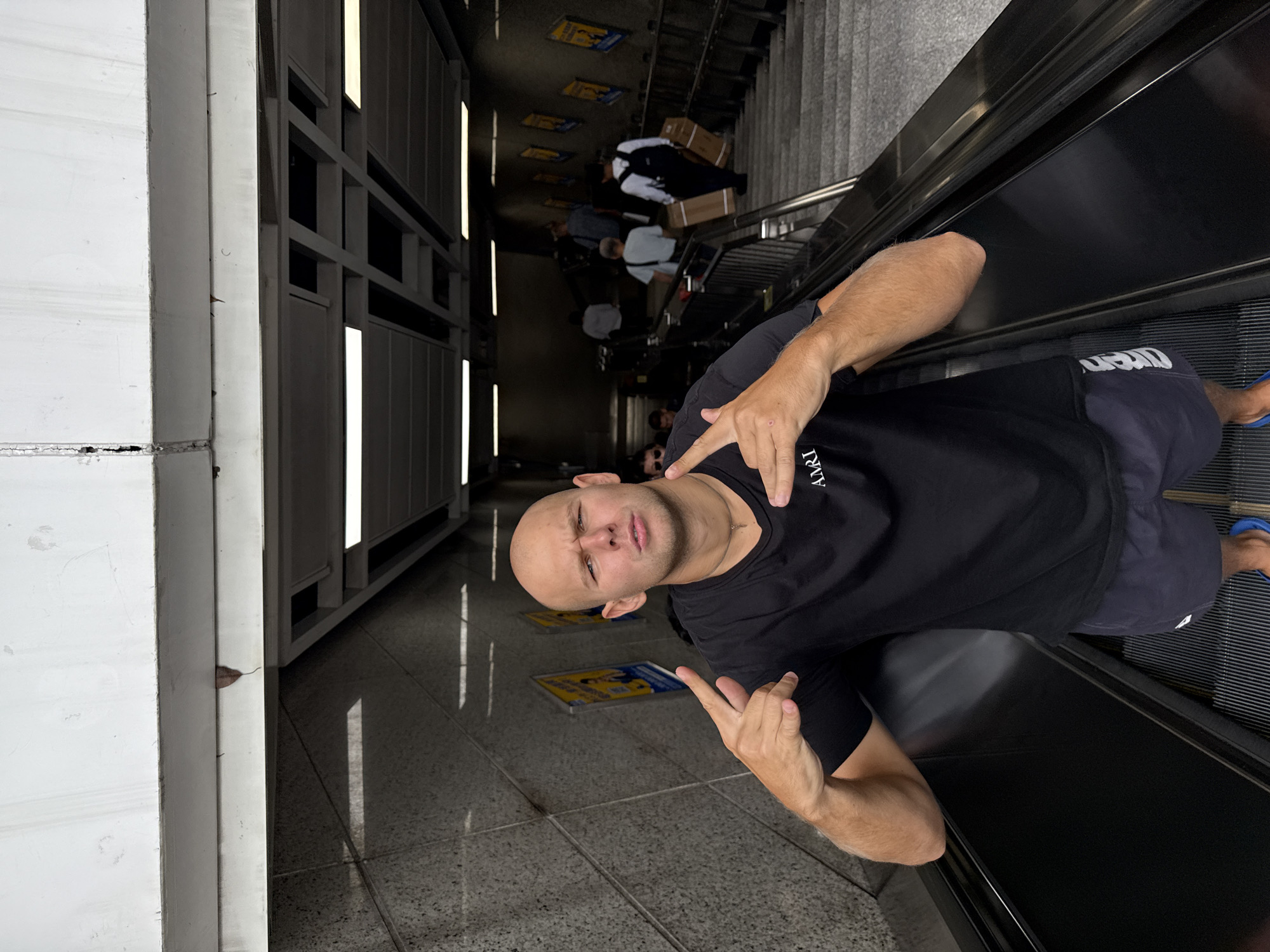 A bald man is standing on a downward moving escalator in what appears to be a subway station in Tianhe District, China. He is wearing a black t-shirt with the logo "AMRI" and dark shorts. He is making a hand gesture with both hands, with his thumbs and index fingers extended in a "rock on" or "peace" sign. The escalator has dark gray treads and a black side. Behind him, other people are seen descending the stairs to the right of the escalator. The walls of the station are made of grey stone-like material, and there are yellow posters visible on the walls. Overhead, a white ceiling with recessed lighting is visible.