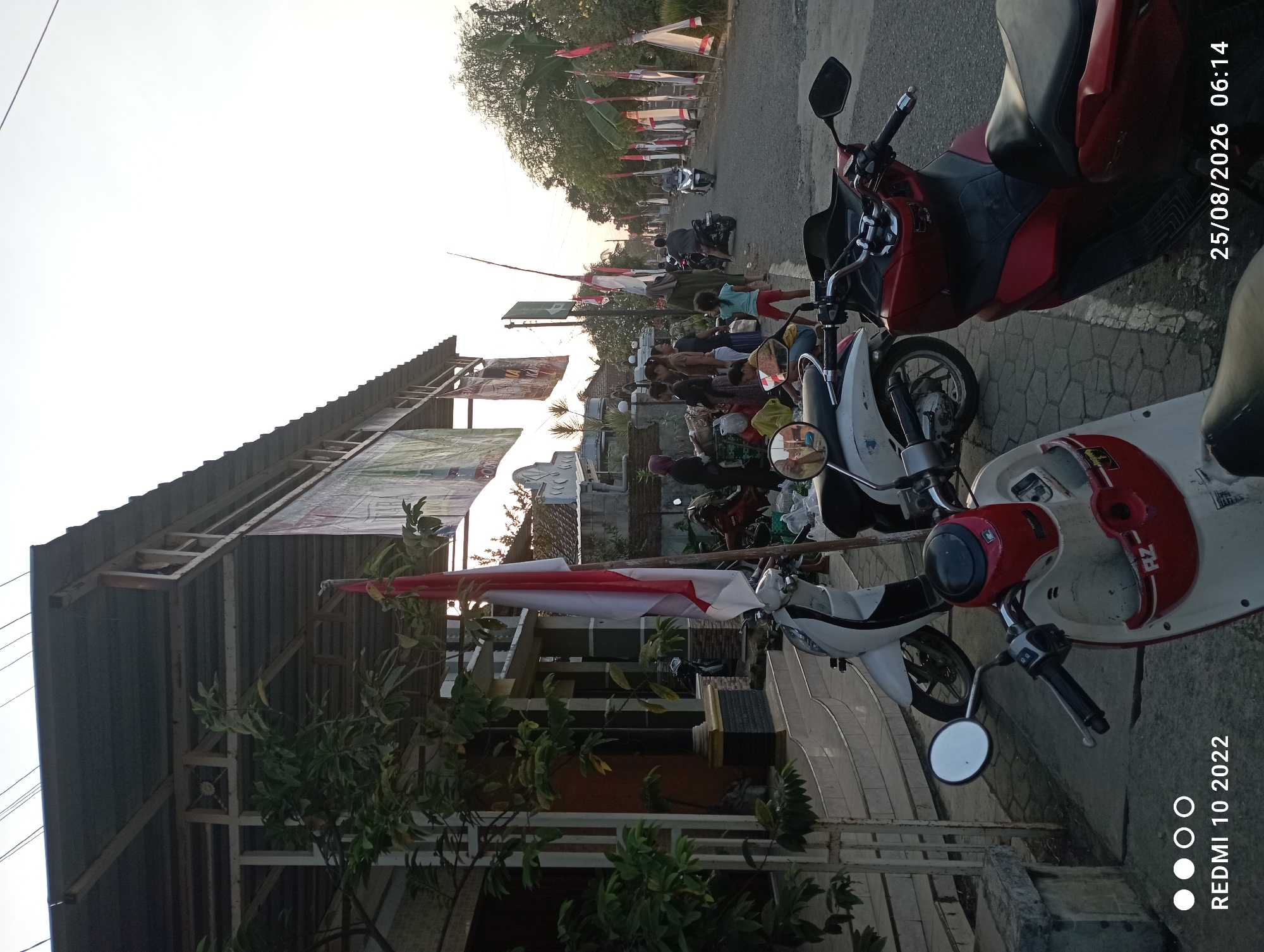 The image depicts a street scene in Dukuhseti, Indonesia, with a focus on the foreground and a road in the background. Several motorcycles are parked on the side of the road, with two prominent in the foreground: a red and black scooter in the lower right and a white scooter with red accents in the lower left. An Indonesian flag, red and white, is planted in the foreground, slightly to the left of center.
In the midground, a group of people are gathered on the side of the road, some near a building with a tin roof supported by metal framing. They appear to be interacting, with some standing and others sitting. A few more motorcycles are visible behind them, and some are in motion on the road.
The road itself is paved and lined with trees on the right side. Along the road, there are numerous small flags, mostly white with some red accents, mounted on poles. The sky is bright, indicating daytime, possibly late afternoon or early morning given the lighting. The overall impression is of a street in a semi-urban or rural Indonesian setting, with a sense of community gathering and local event preparation.