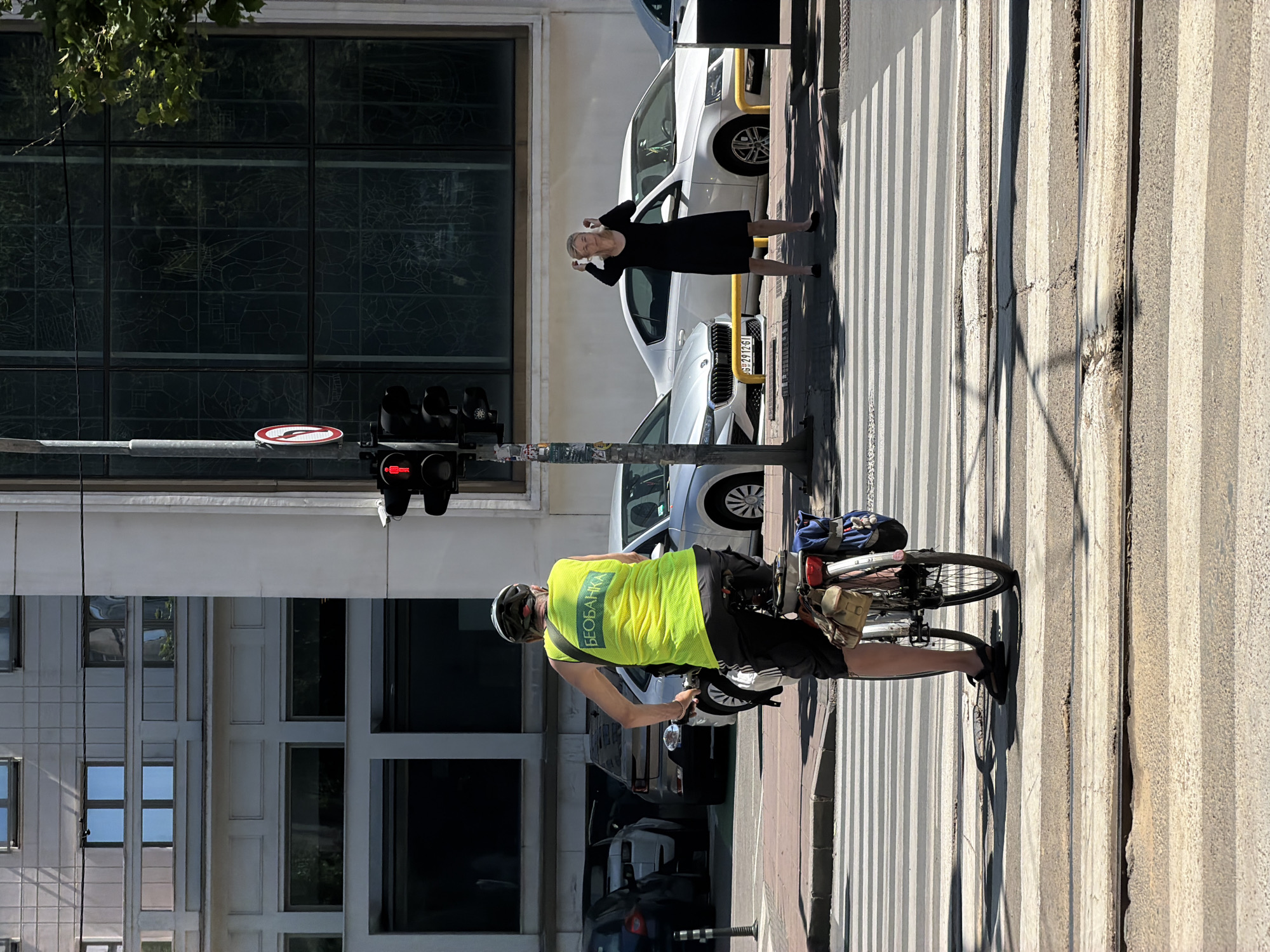 An individual wearing a reflective yellow vest with Cyrillic lettering "БЕОБАНКА" rides a bicycle across a crosswalk in Vracar Urban Municipality, Serbia. The cyclist, viewed from behind, is wearing a helmet and dark shorts, with bags attached to the rear of the bicycle. To the right, a woman in a black dress stands on the sidewalk, holding her hands to her head. Behind them, several cars are parked or in motion. A traffic light shows a red pedestrian signal. The background features a modern building with large windows. The scene is set on a sunny day, with shadows cast across the crosswalk.