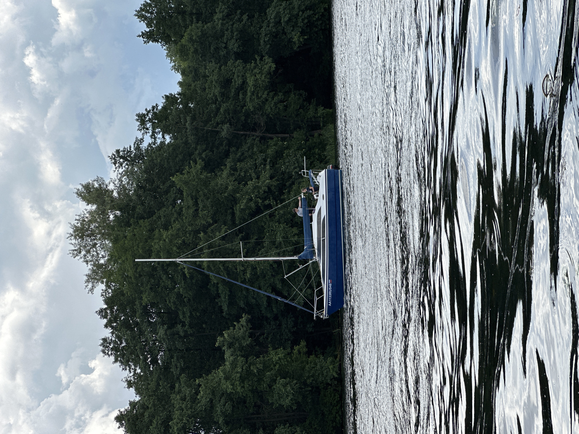 A blue sailboat with white trim is on the water near a dense line of dark green trees. The sailboat has a mast and rigging. Two people are visible on the deck of the sailboat. The water is choppy with reflections of the sky and trees. The sky is cloudy with patches of blue. The sailboat has the registration "AA1133RUS48" on its side. The scene is likely a body of water in or near Lipetsk, Russia, given the "RUS48" designation which typically indicates the Lipetsk Oblast.