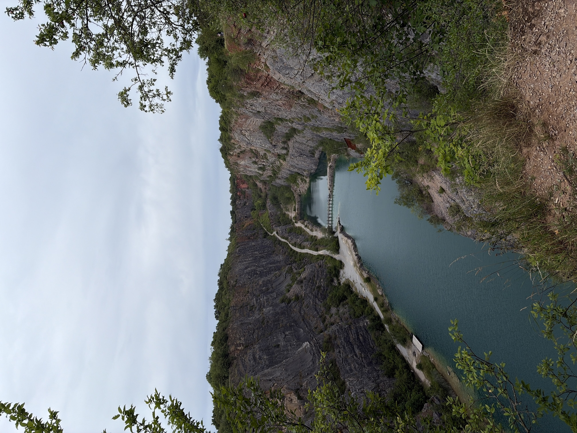 The image shows a quarry filled with turquoise water in Trněný Újezd, Czechia. Steep, rocky cliffs surround the water, with a path winding along the edge. A small wooden dock is visible on the left side of the water. In the distance, a bridge crosses the quarry. Green trees and vegetation grow along the tops of the cliffs and on the slopes. The sky is overcast.