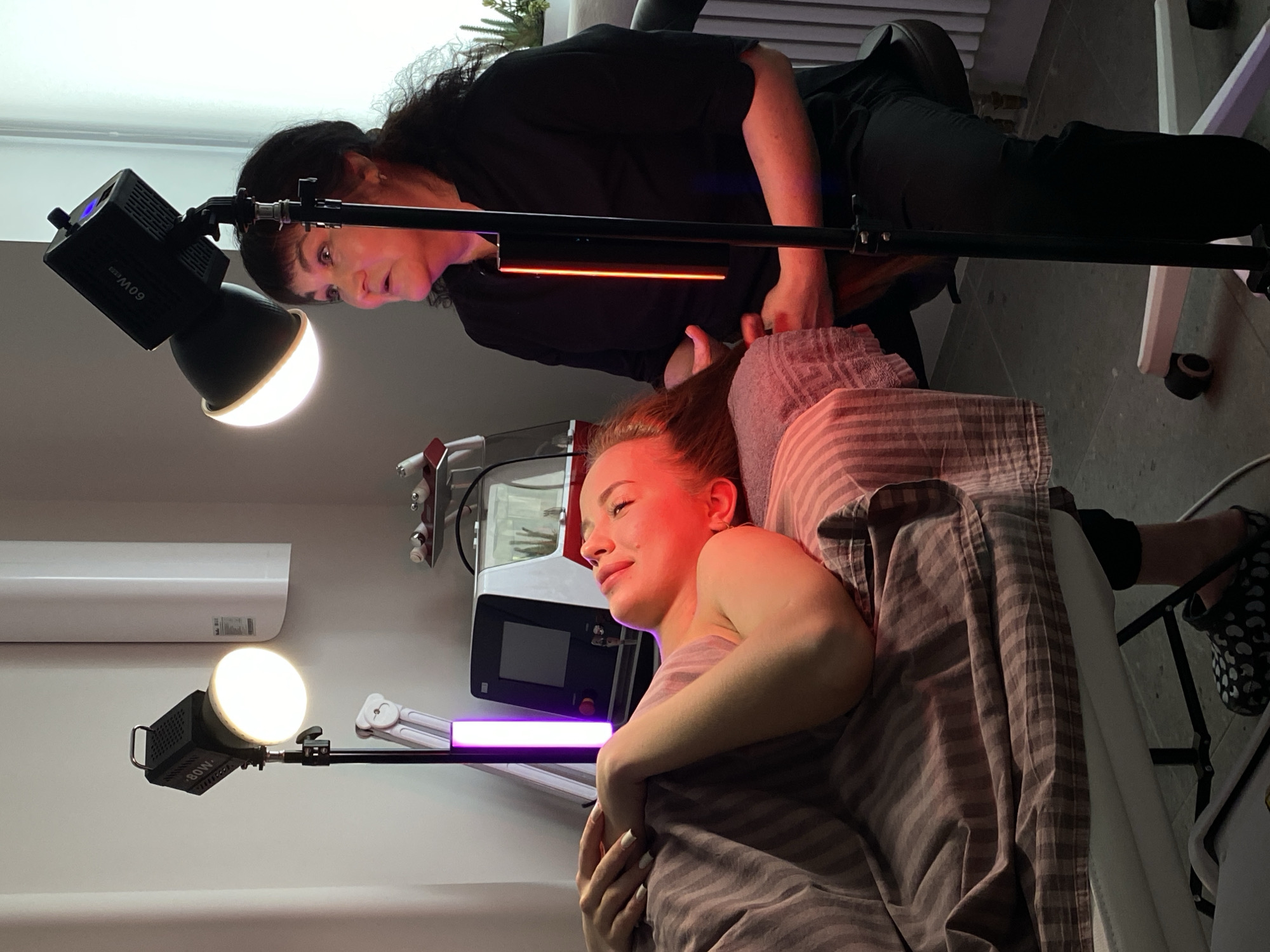 A woman lies covered with a striped blanket on a treatment bed, receiving a beauty treatment at what appears to be a salon or spa. An older woman with dark hair is attending to her, possibly applying a product or performing a procedure. Professional lighting equipment, including what looks like a 60W bulb on a stand and a rectangular light bar, surrounds the scene. There is a white medical device visible next to the patient. An air conditioner is mounted high on the wall. The location is possibly in Izhevsk, Russia.