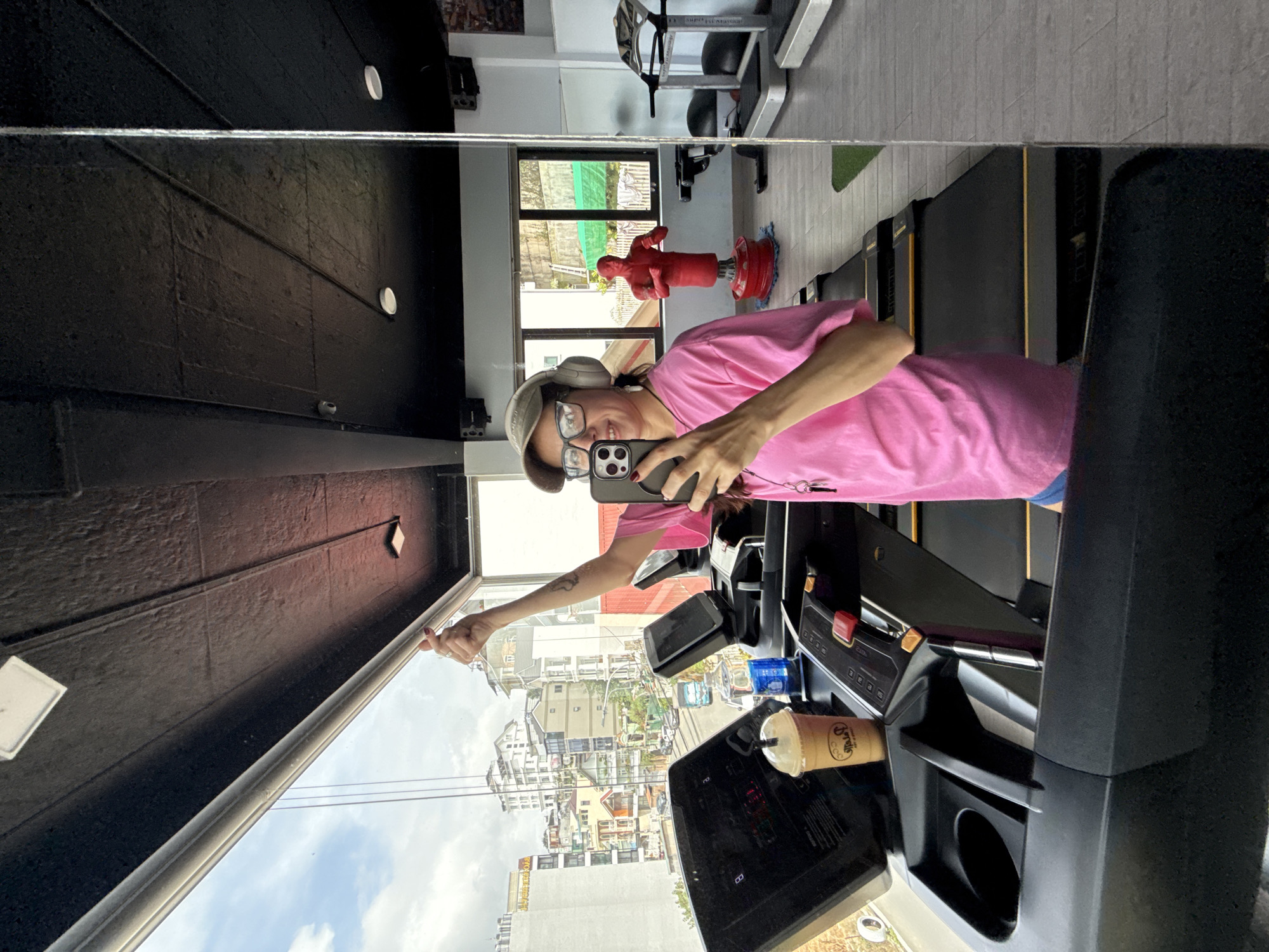 A person is taking a selfie in a gym with a large window overlooking a cityscape. The person is wearing a pink t-shirt, blue shorts, a baseball cap, glasses, and headphones. They are holding a smartphone in their right hand and giving a thumbs-up with their left hand. In the foreground, a treadmill is visible, with a cup of coffee on it. In the background, a red punching dummy and other gym equipment are present. The cityscape outside the window shows buildings and a street with vehicles, suggesting an urban setting in Da Lat, Vietnam. The sky is cloudy.