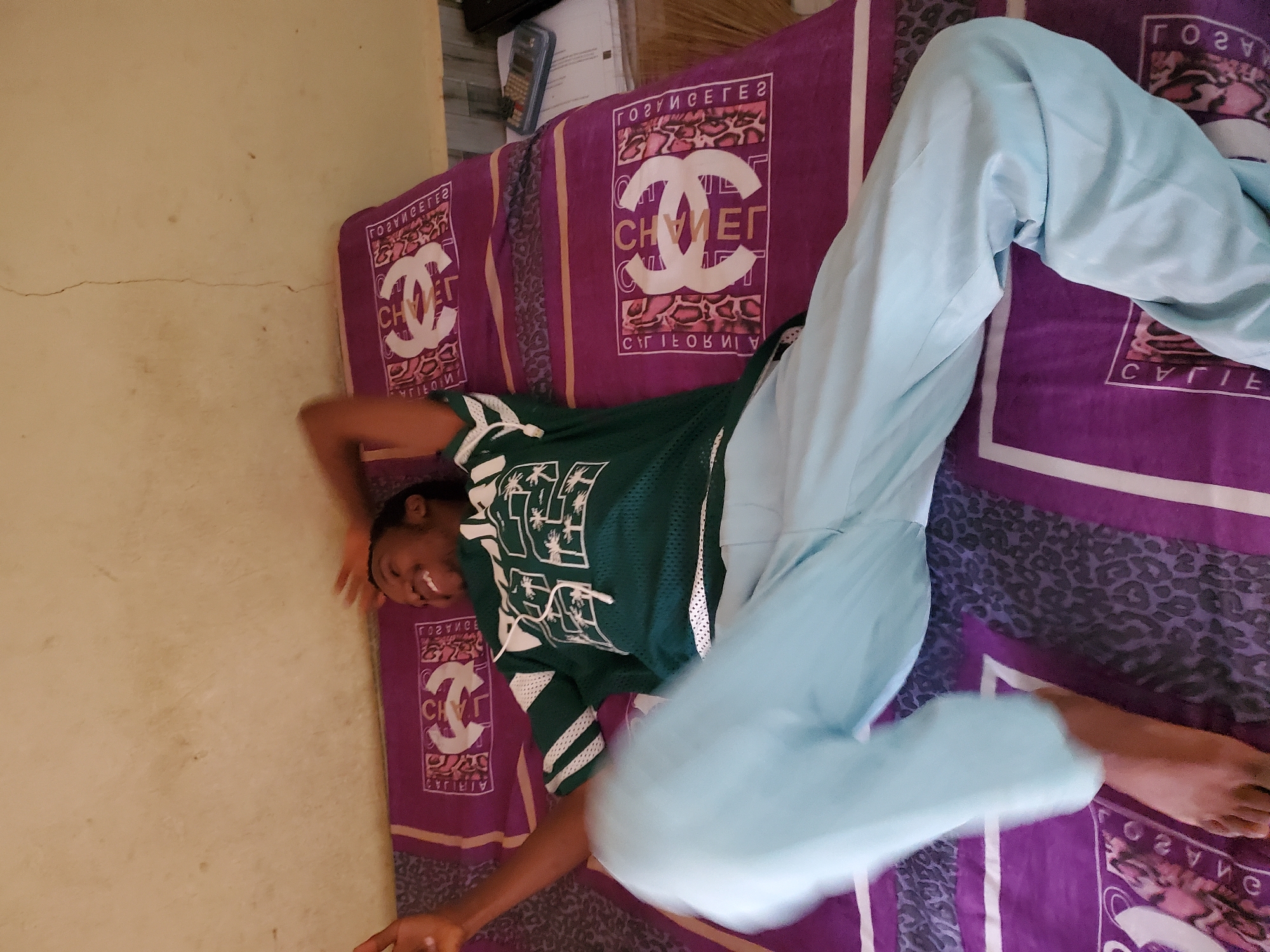 A young Black male is lying on a bed, propped up on his elbows, with his legs extended and bent. He is wearing a green jersey with white trim and a white graphic print, and light blue pants. His eyes are closed, and he has a wide smile on his face. The bed is covered with a purple and black patterned bedsheet. The wall behind the bed is a light brown color with some stains and a visible crack. To the right of the bed, a stack of papers and a calculator are visible. The scene is likely indoors, possibly in a bedroom, and given the context of the location being provided as Jalingo, Nigeria, it is a domestic setting within the city.